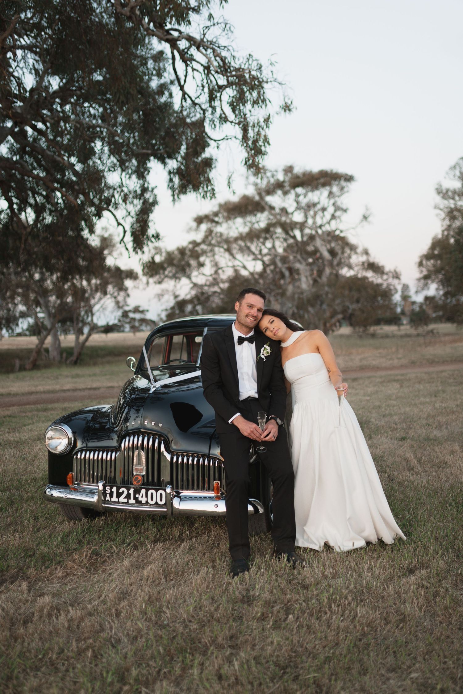 Bride and groom leaning against a vintage black Holden car in a rustic field at dusk, surrounded by eucalyptus trees.