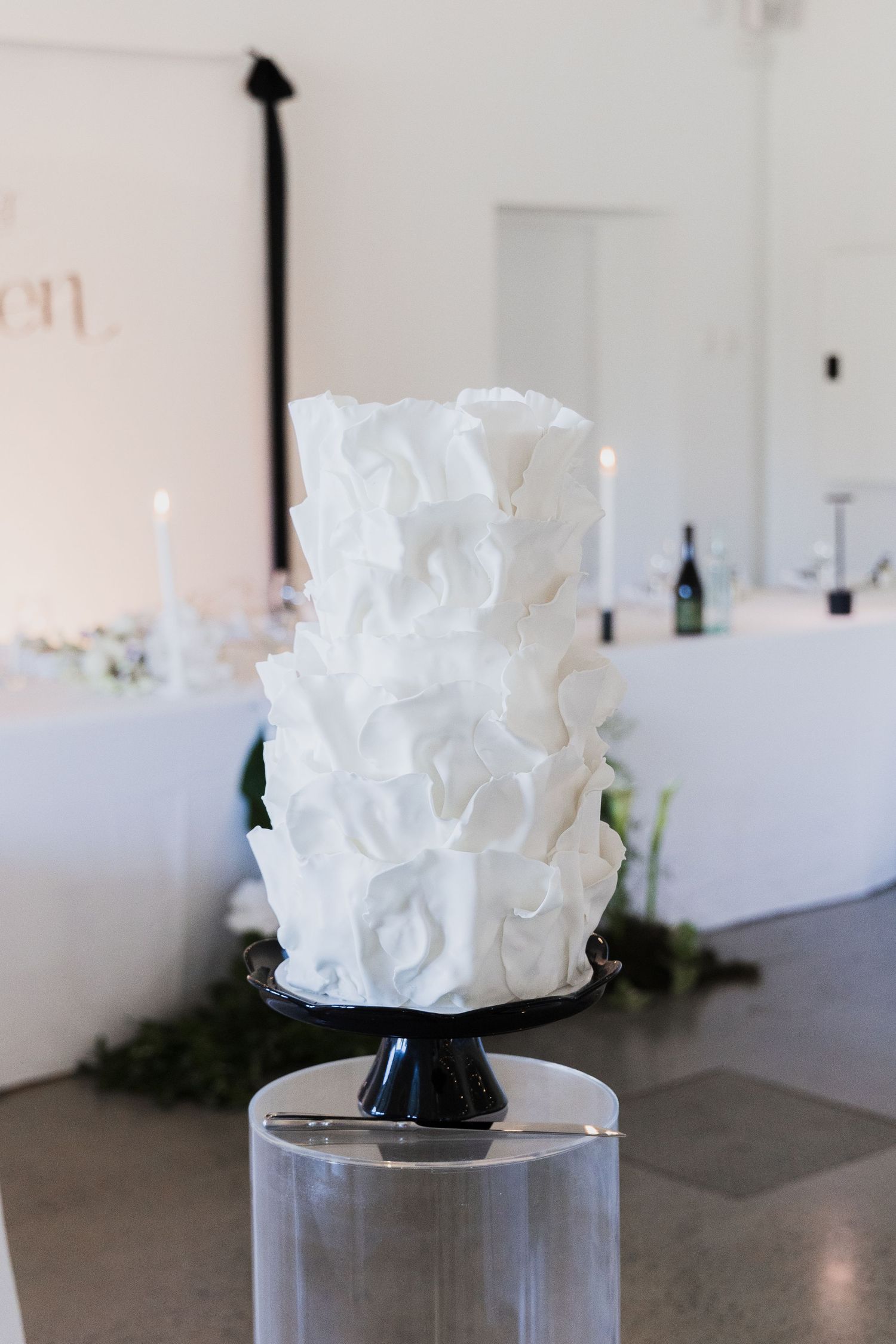 An elegant white textured wedding cake on a clear pedestal against a minimalist white background.