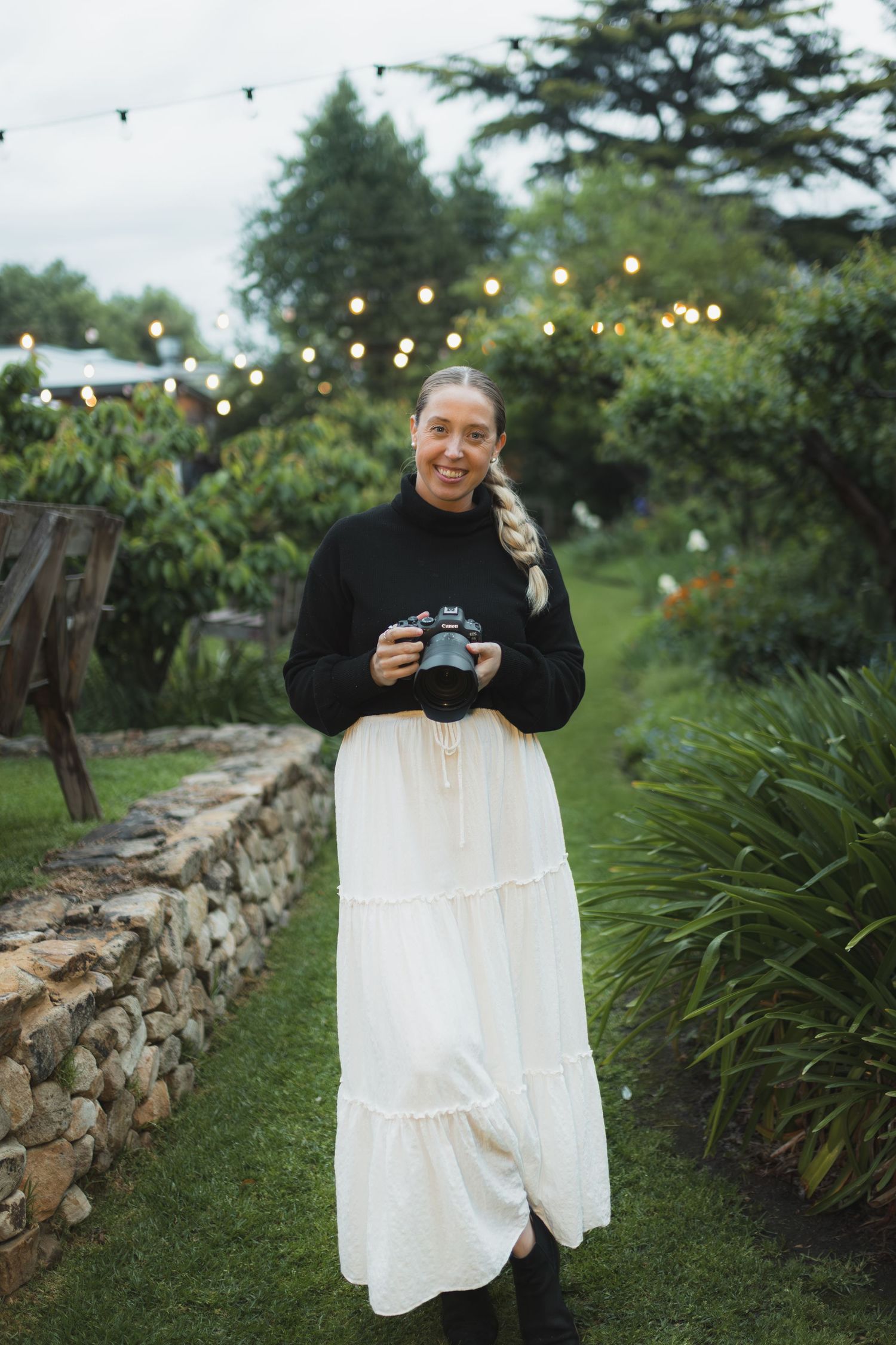 Woman in black sweater and white maxi skirt holds camera in lush garden with string lights.