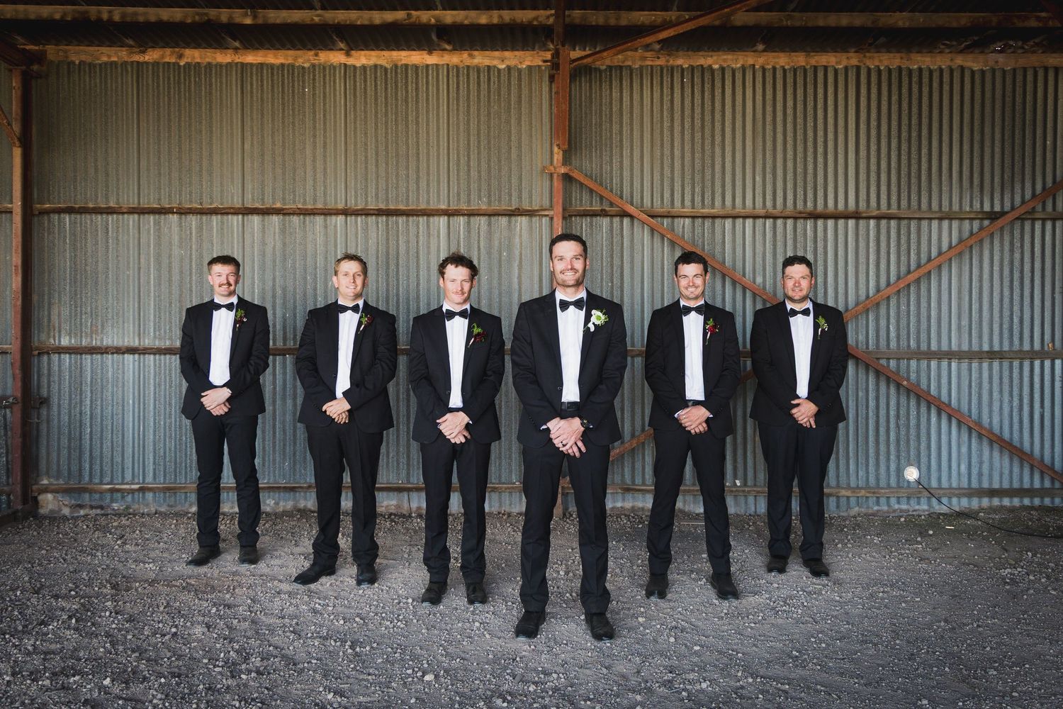 Six groomsmen in black tuxedos and bow ties pose in a rustic barn for a wedding party photo.