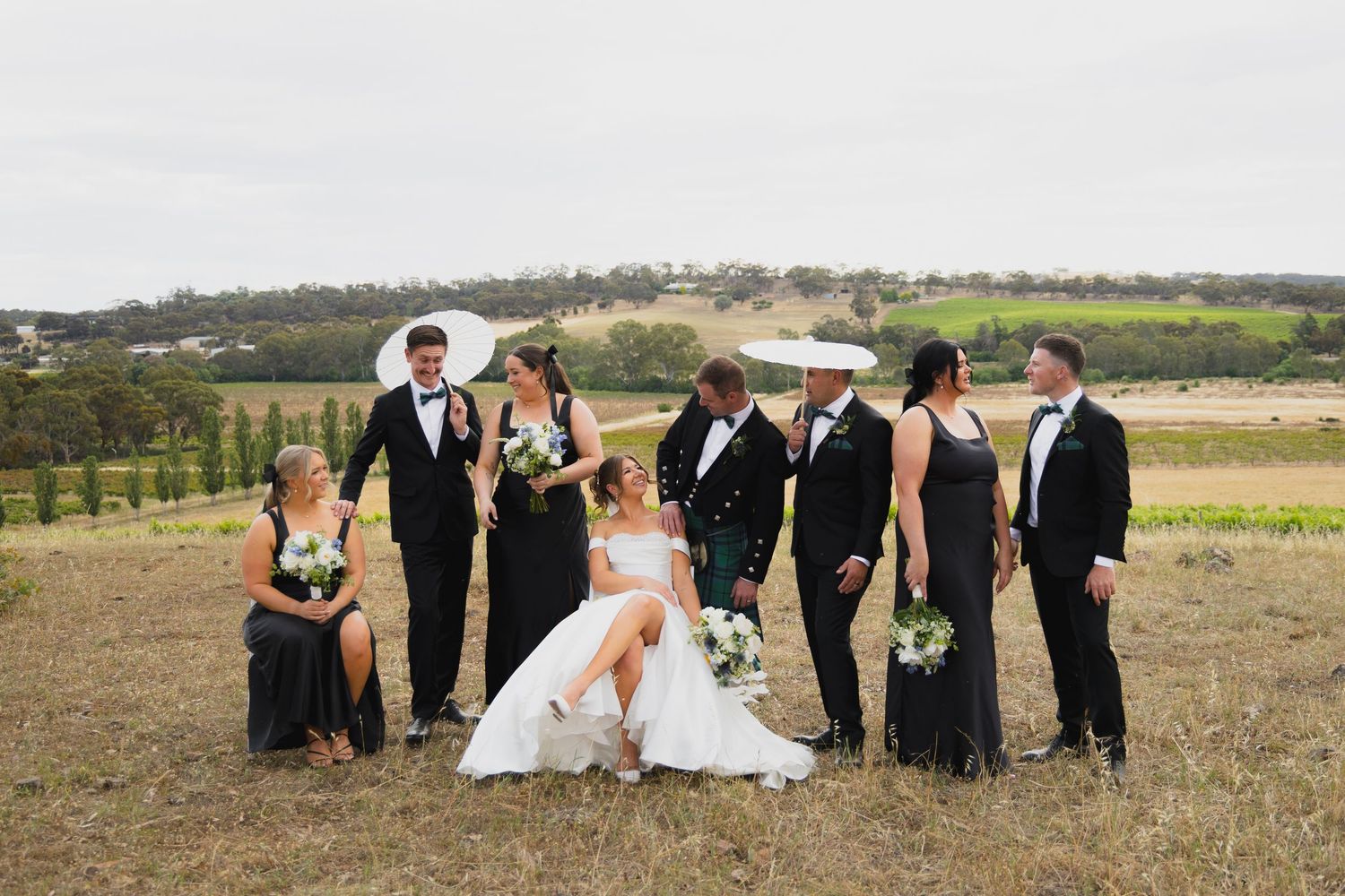 Wedding party in black formal wear and kilts pose in a vineyard field with rolling hills in the background.