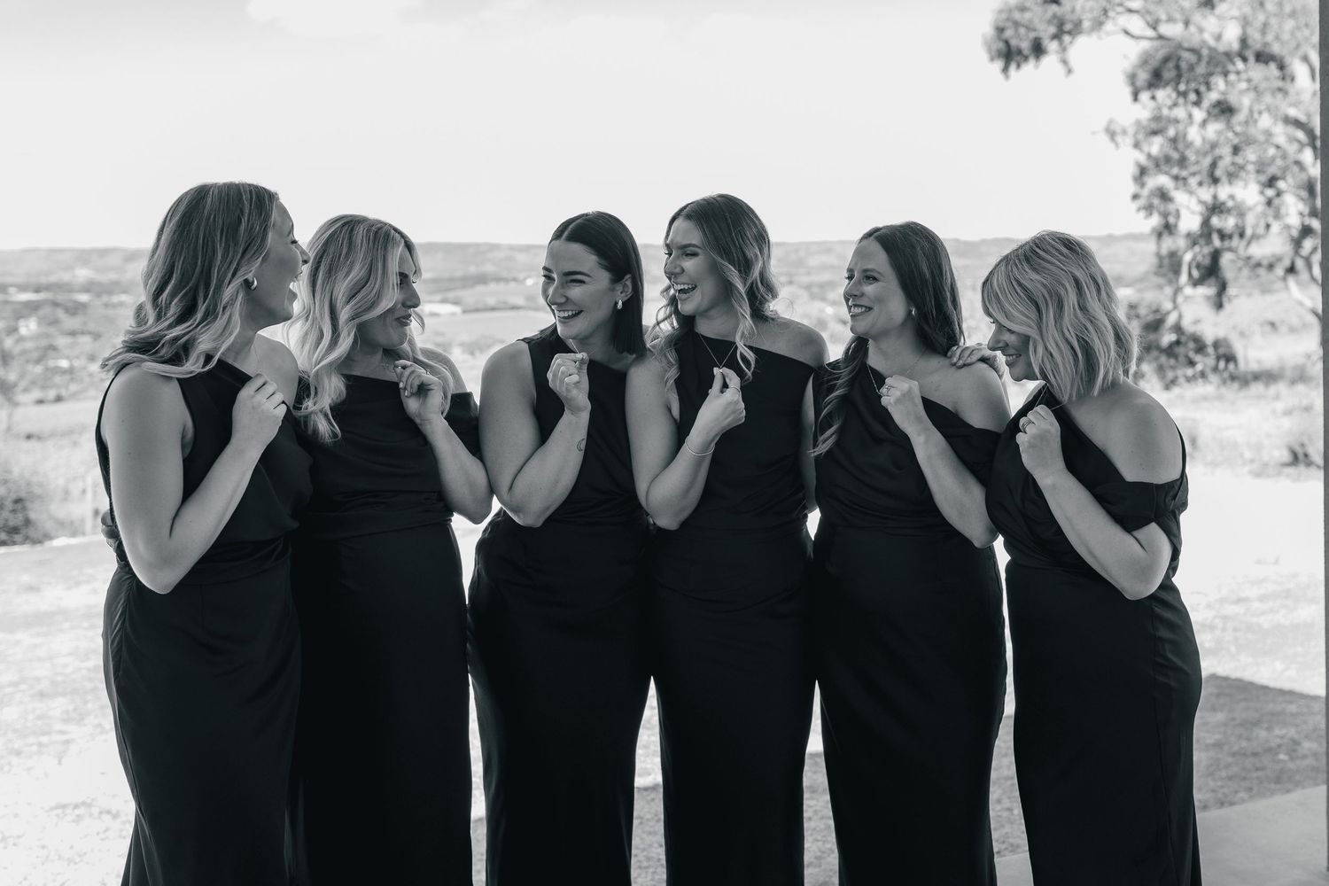 A group of six friends in matching black dresses smile and laugh together in a black and white photograph.