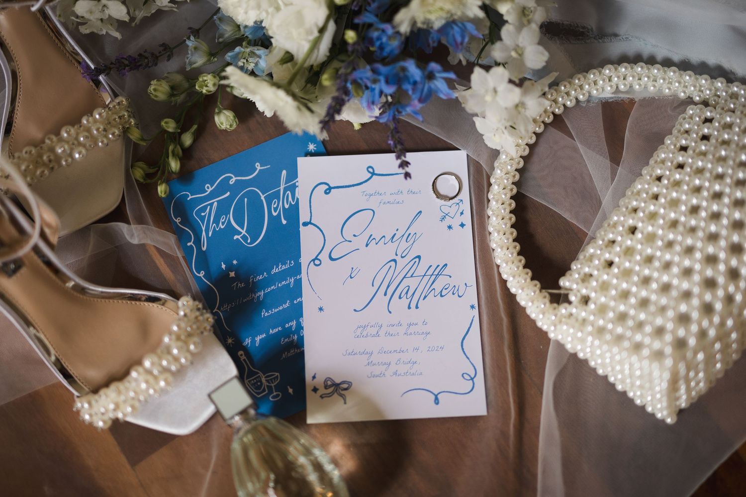 Wedding invitation card surrounded by blue flowers, pearl beadwork accessories, and elegant styling elements.
