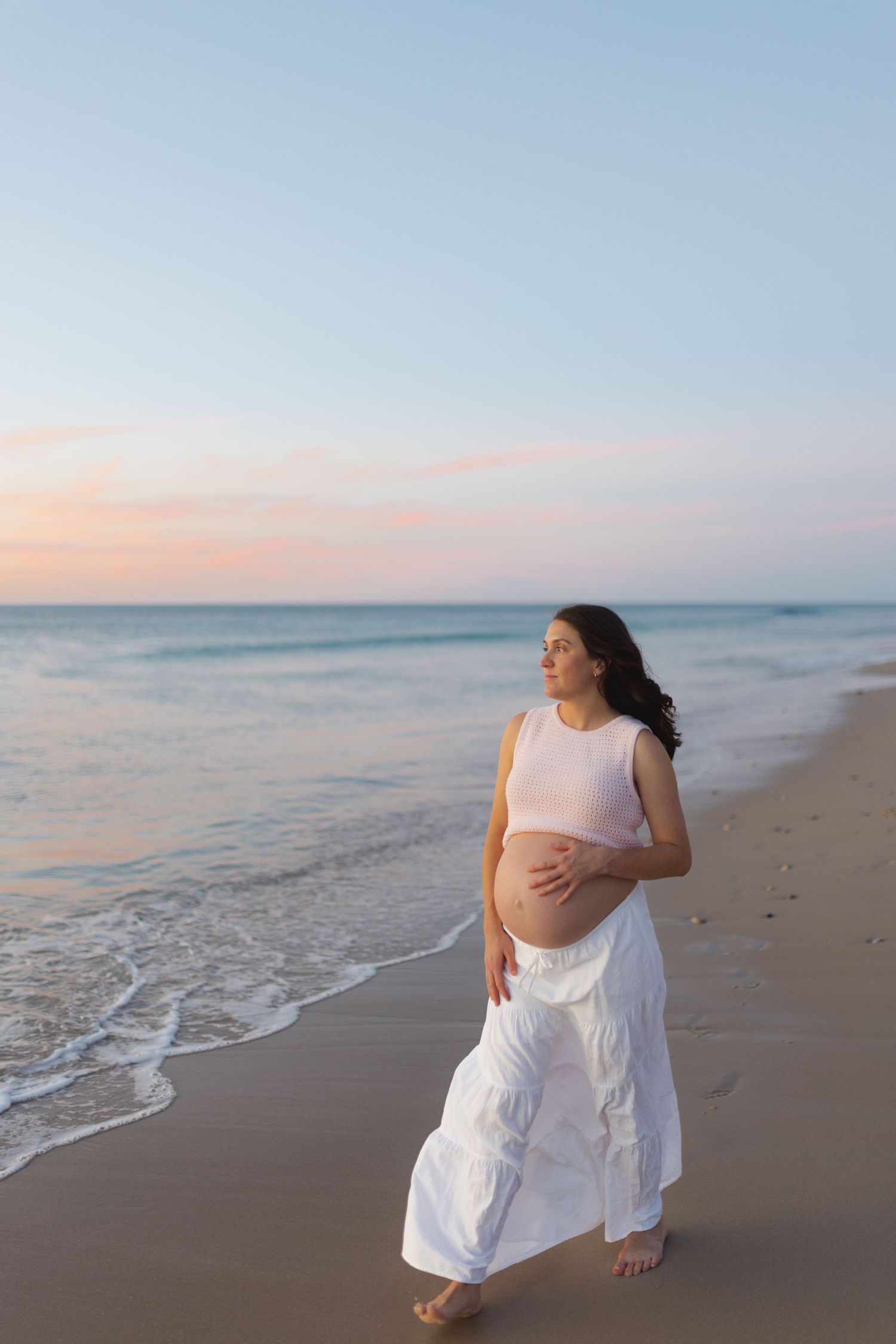 An expectant mother in white dress walks along peaceful beach at sunset.