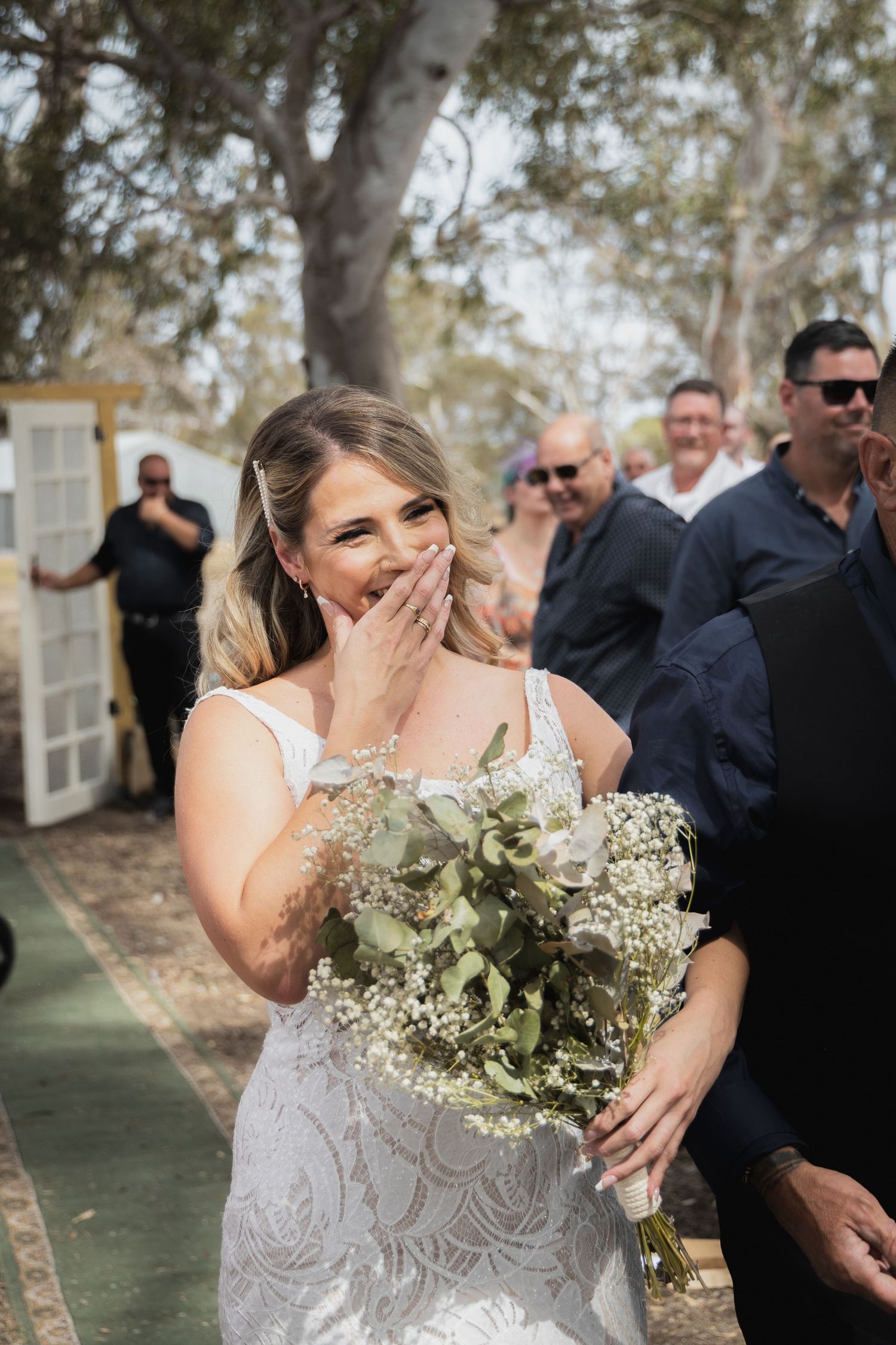 A bride in a lace wedding dress holds a white and green bouquet while expressing joy during an outdoor ceremony.
