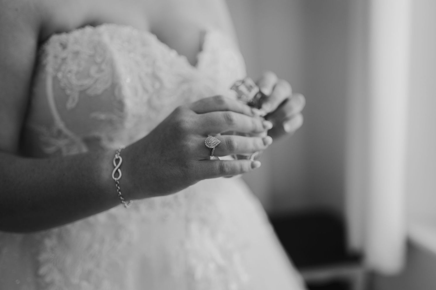Black and white photo showing hands wearing jewelry against a white lace dress with soft natural lighting.