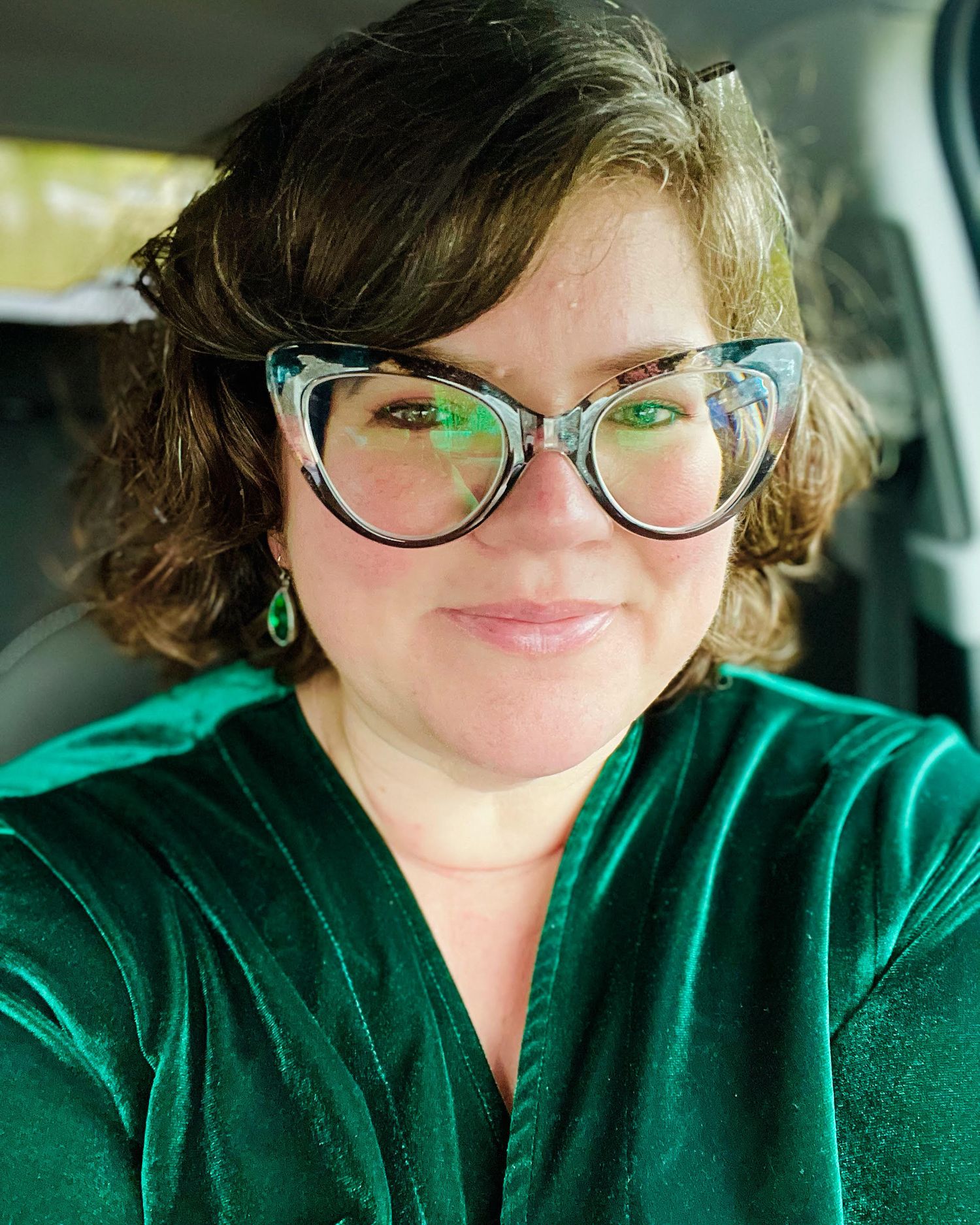 Someone wearing cat-eye glasses and a green velvet top takes a selfie in a car.