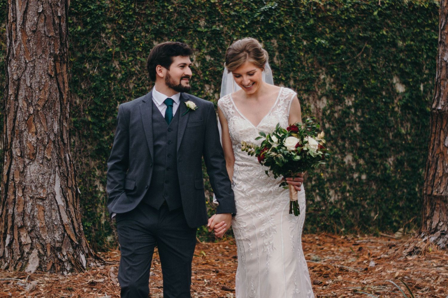 Couple walks together on rustic forest path during autumn wedding ceremony with bride holding white floral bouquet.