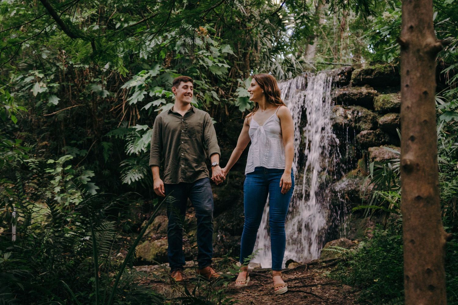 A couple holds hands while standing on a forest path near a cascading waterfall in a lush natural setting.
