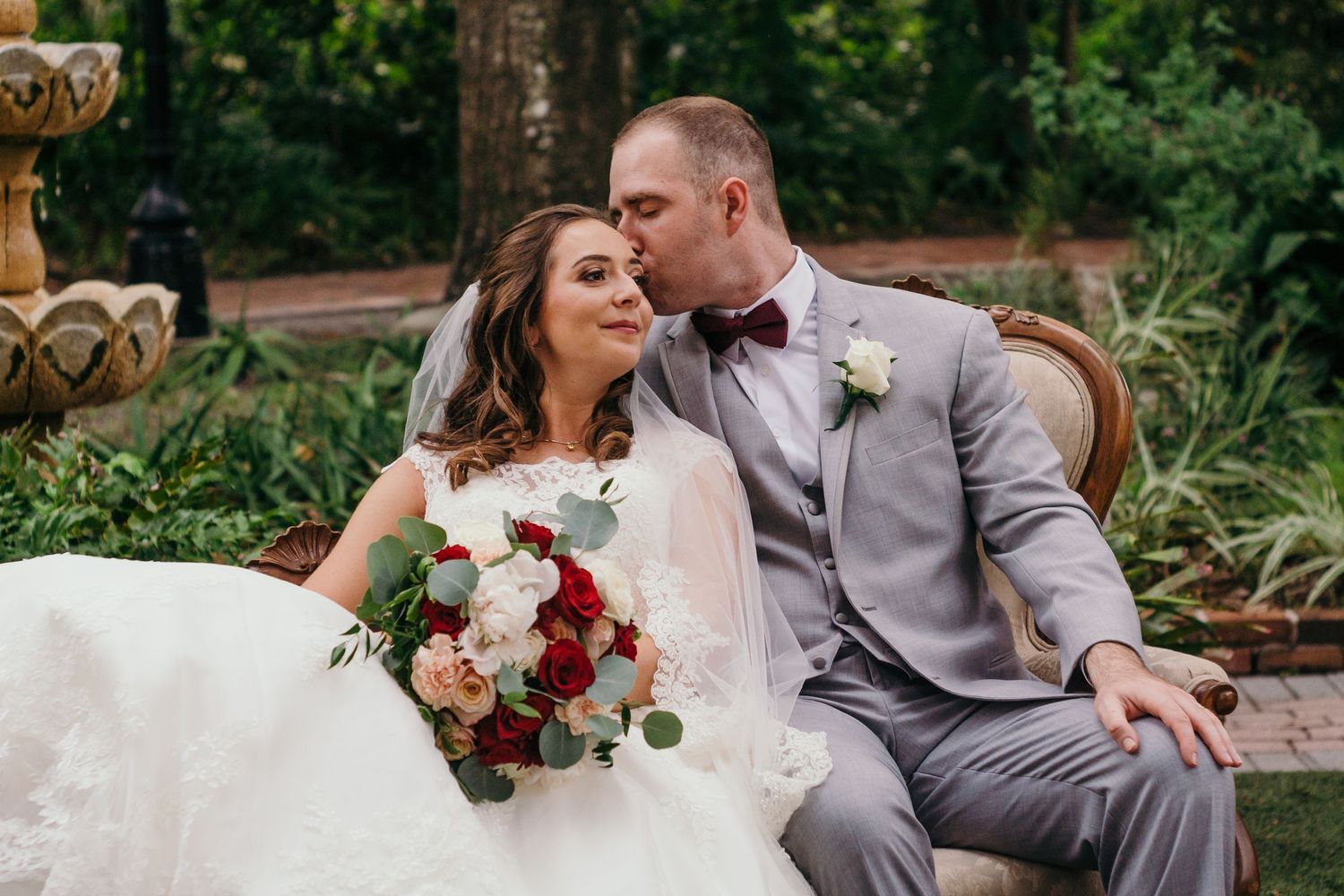 A romantic garden wedding scene with a couple in formal attire sharing an intimate moment on an ornate bench.