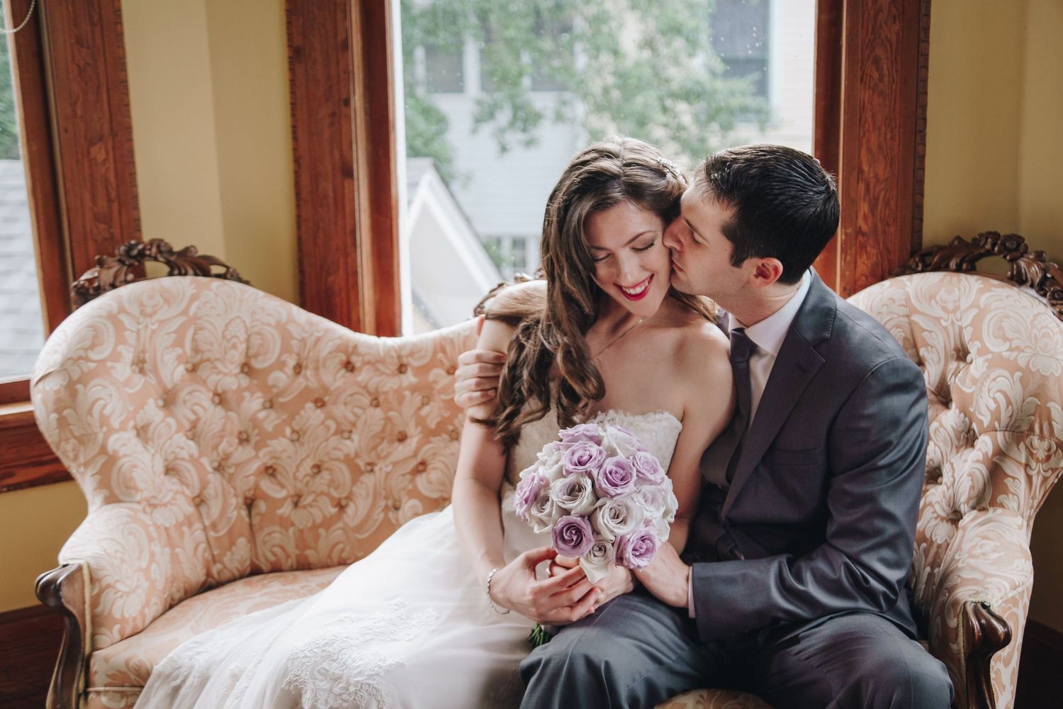 Romantic wedding couple sits close together on vintage pink tufted loveseat holding floral bouquet by window.