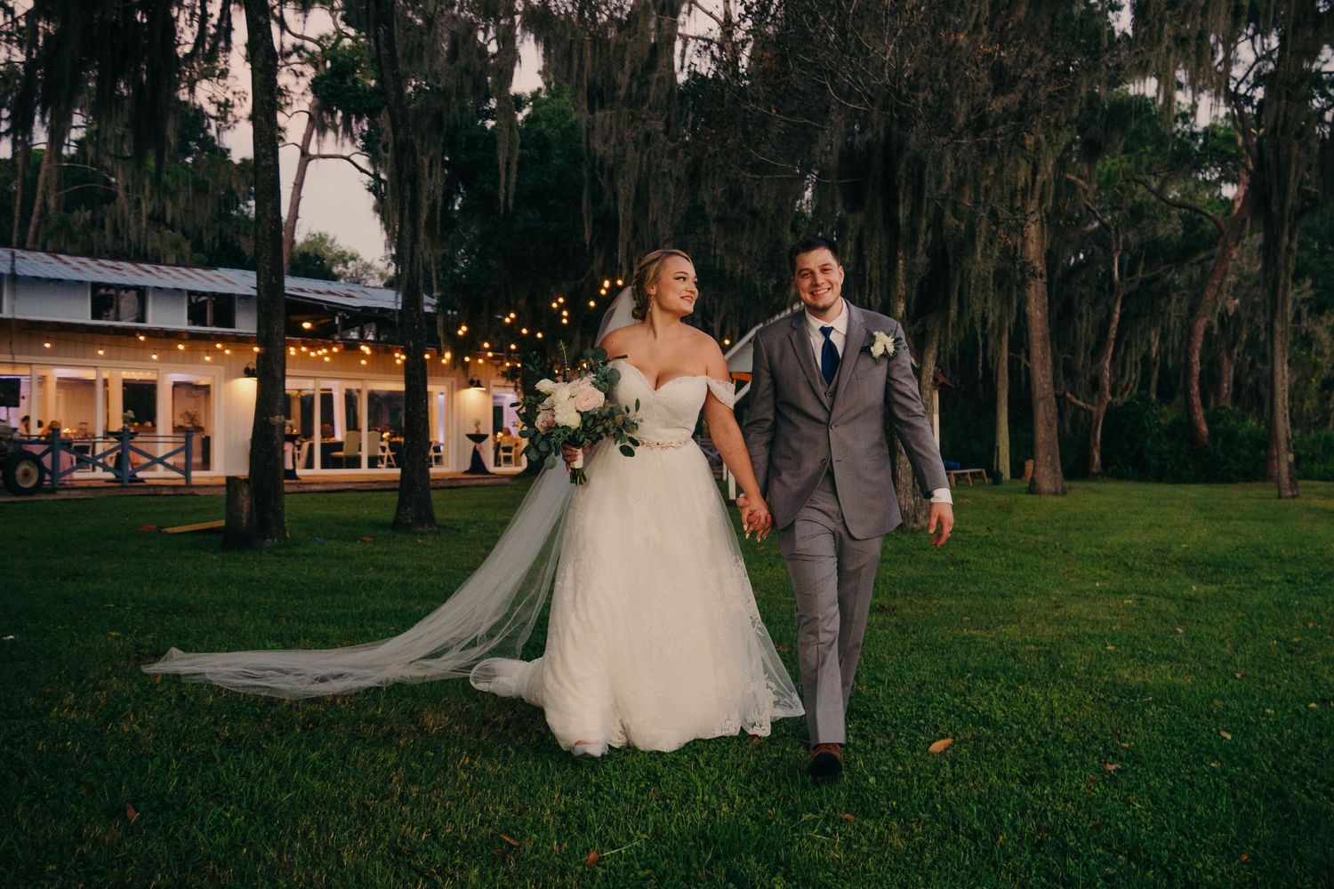 A couple in wedding attire walks together on a green lawn surrounded by trees at sunset. Tampa, Gainesville, Orlando wedding photographer