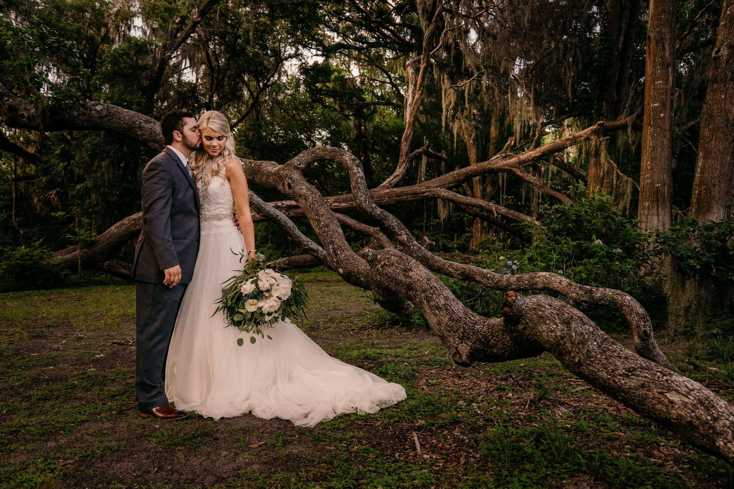 Wedding couple stands together near a twisted tree trunk in a romantic forest setting at dusk.