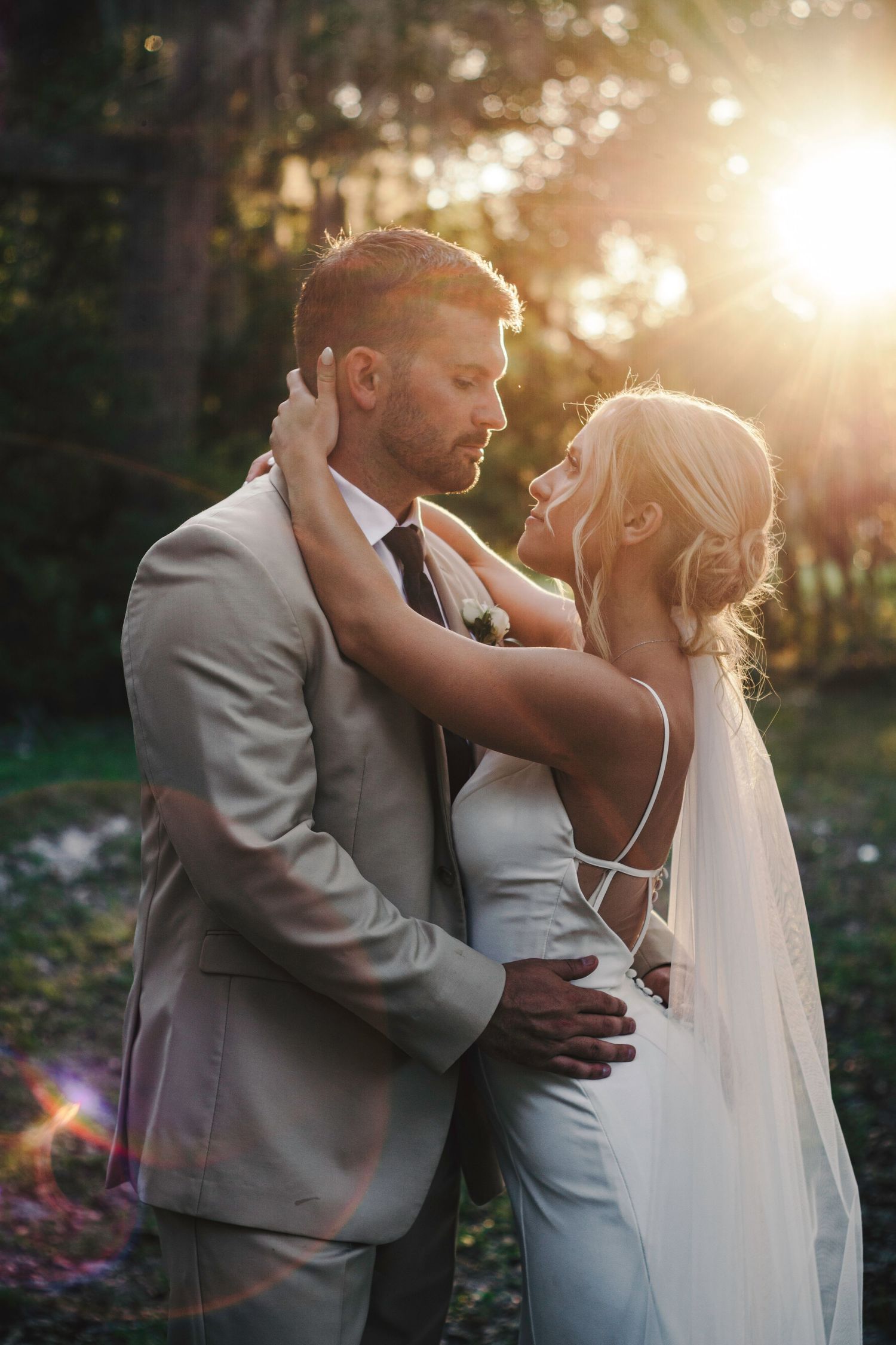 A romantic wedding couple embraces in golden sunlight during sunset, with the bride wearing a flowing veil and white dress.
