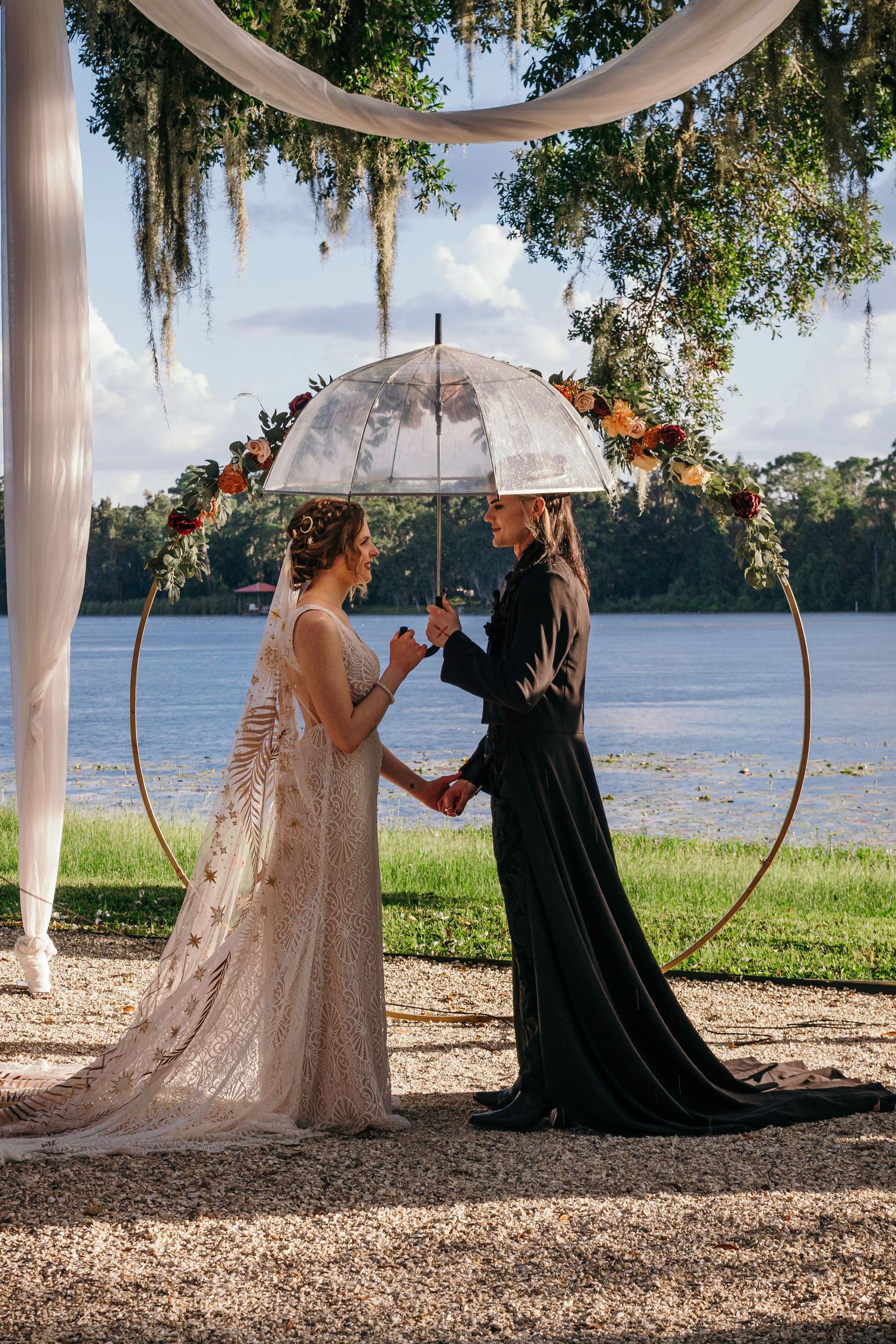 A romantic outdoor wedding ceremony by a lake with a decorative circular arch and translucent umbrella.