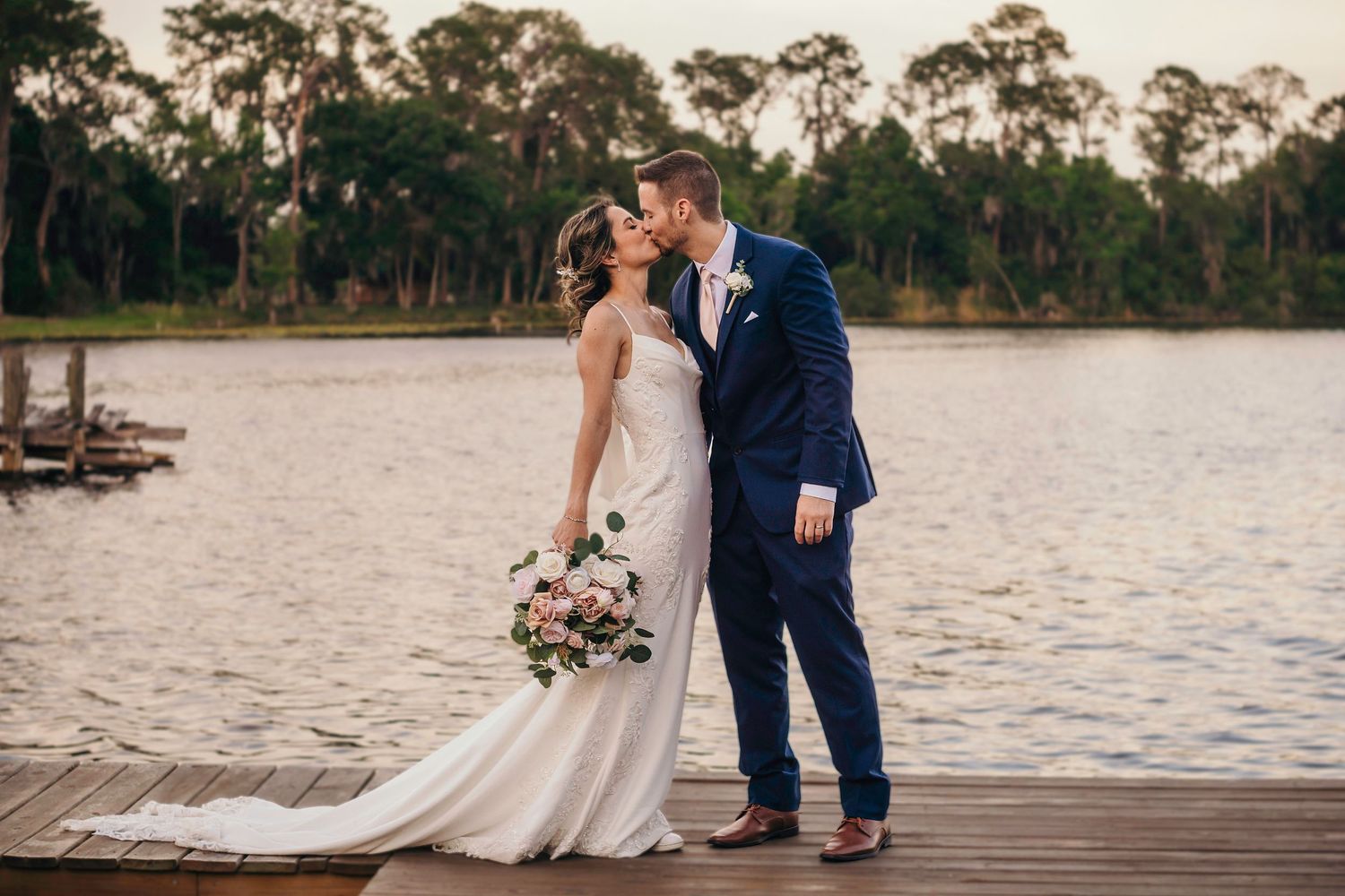A romantic couple shares a tender moment on a wooden dock by a serene lake at sunset, with a beautiful bouquet in hand.