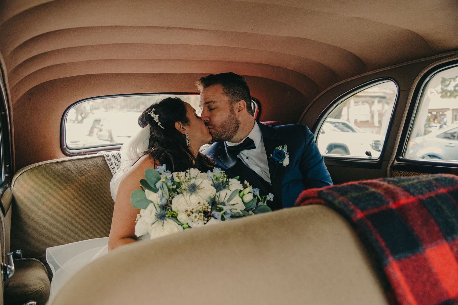 Newlywed couple sharing an intimate moment in a vintage car with white floral bouquet and red plaid blanket.