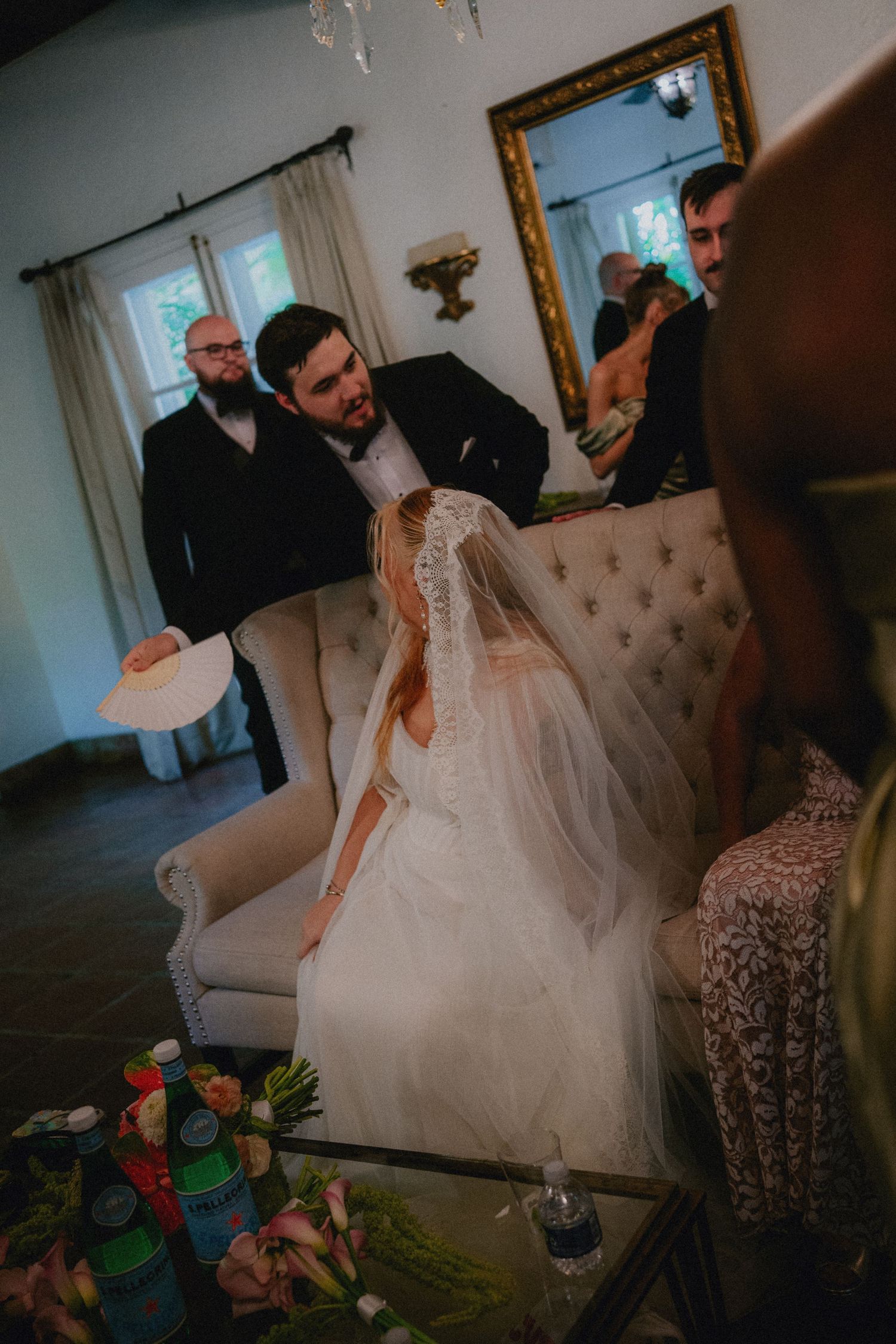 Bride in white lace gown and veil sits on tufted sofa as groom in black suit leans over her indoors.
