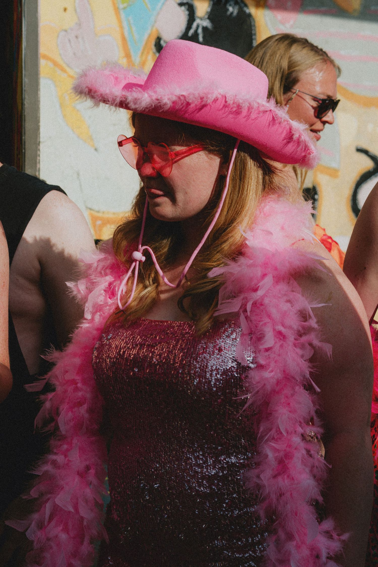 Woman in pink sequin dress, feather boa, cowboy hat, and heart glasses at a festive outdoor event.