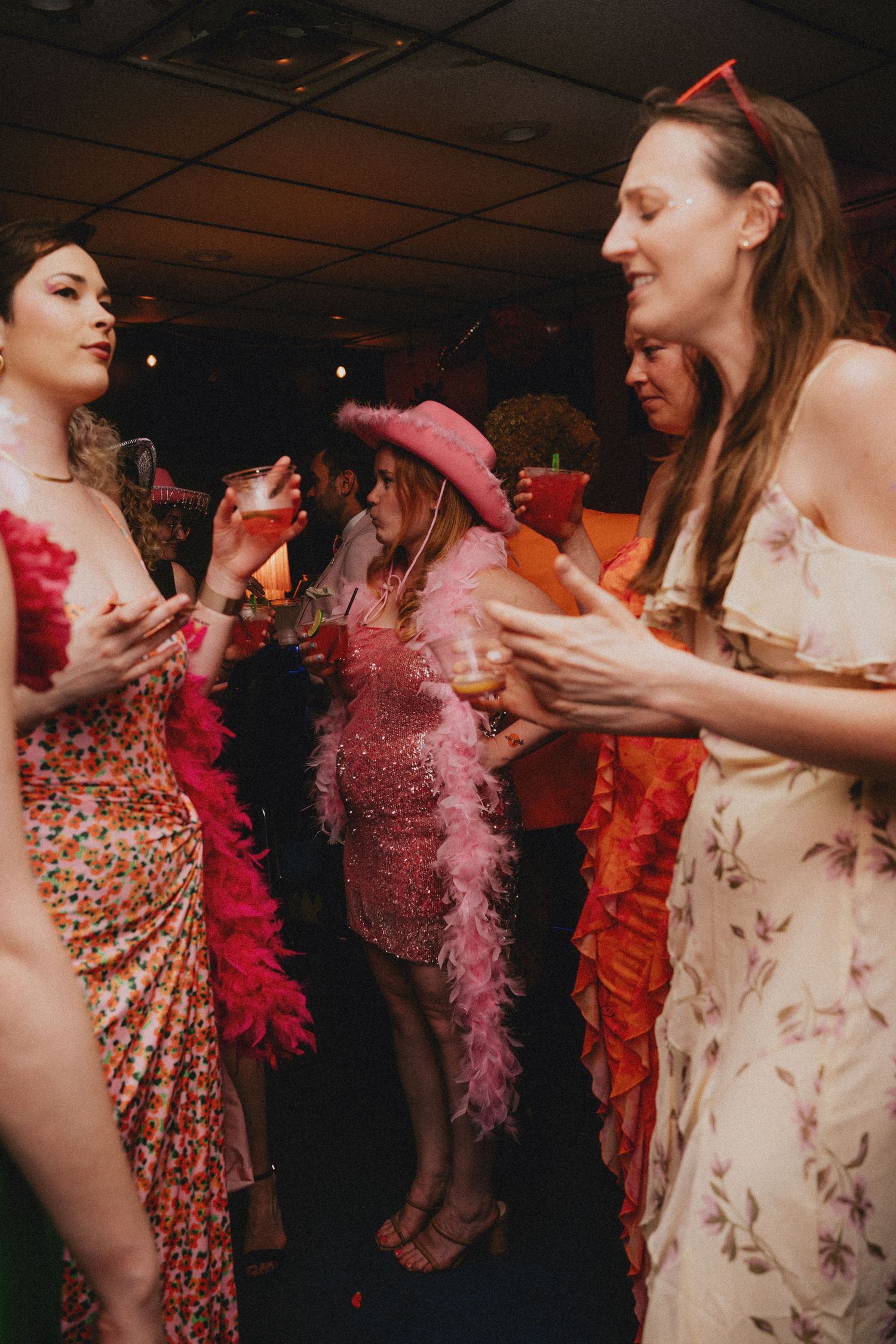 Women in colorful party dresses and pink feather boas dancing and socializing at a festive indoor event.