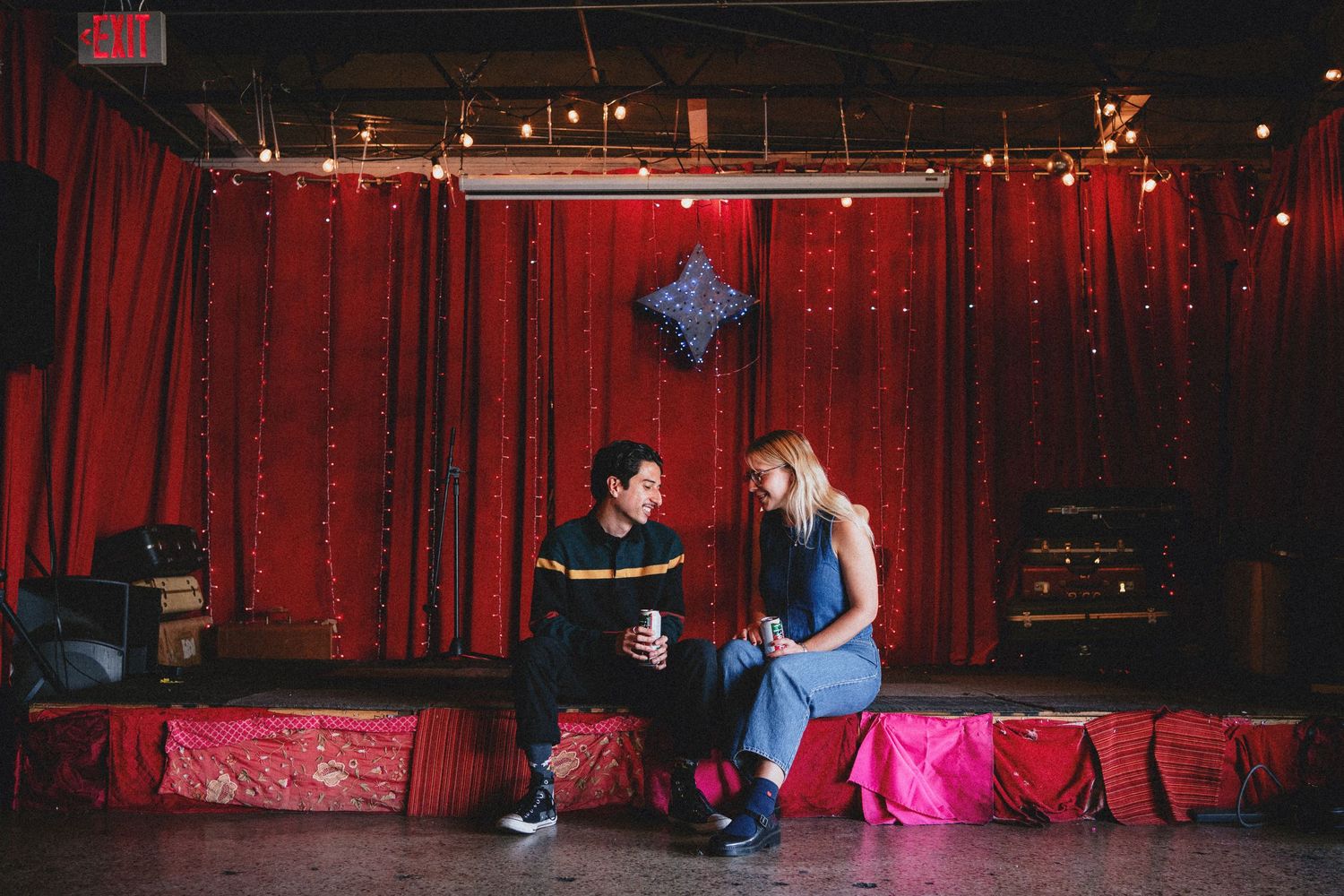 A man and woman sit together on a stage with red curtains and string lights in a cozy venue.