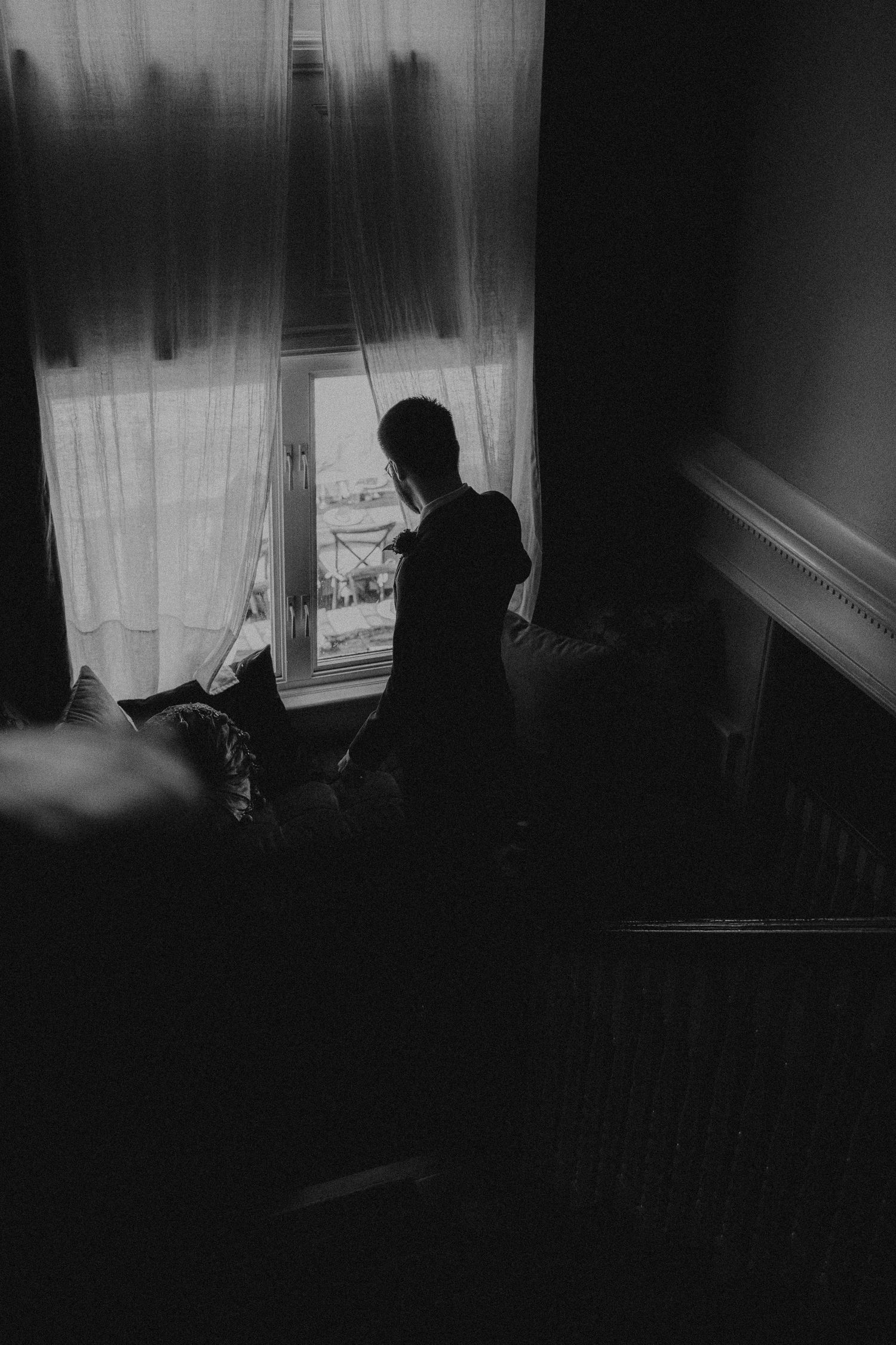 Silhouette of a person standing by a curtained window in a dark moody room, black and white photography.