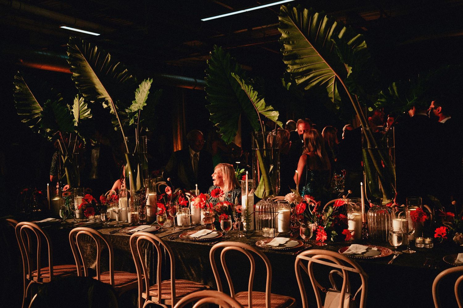 Dramatic tropical wedding reception with dark ambiance, red floral centerpieces, candles, and lush banana leaf arrangements.