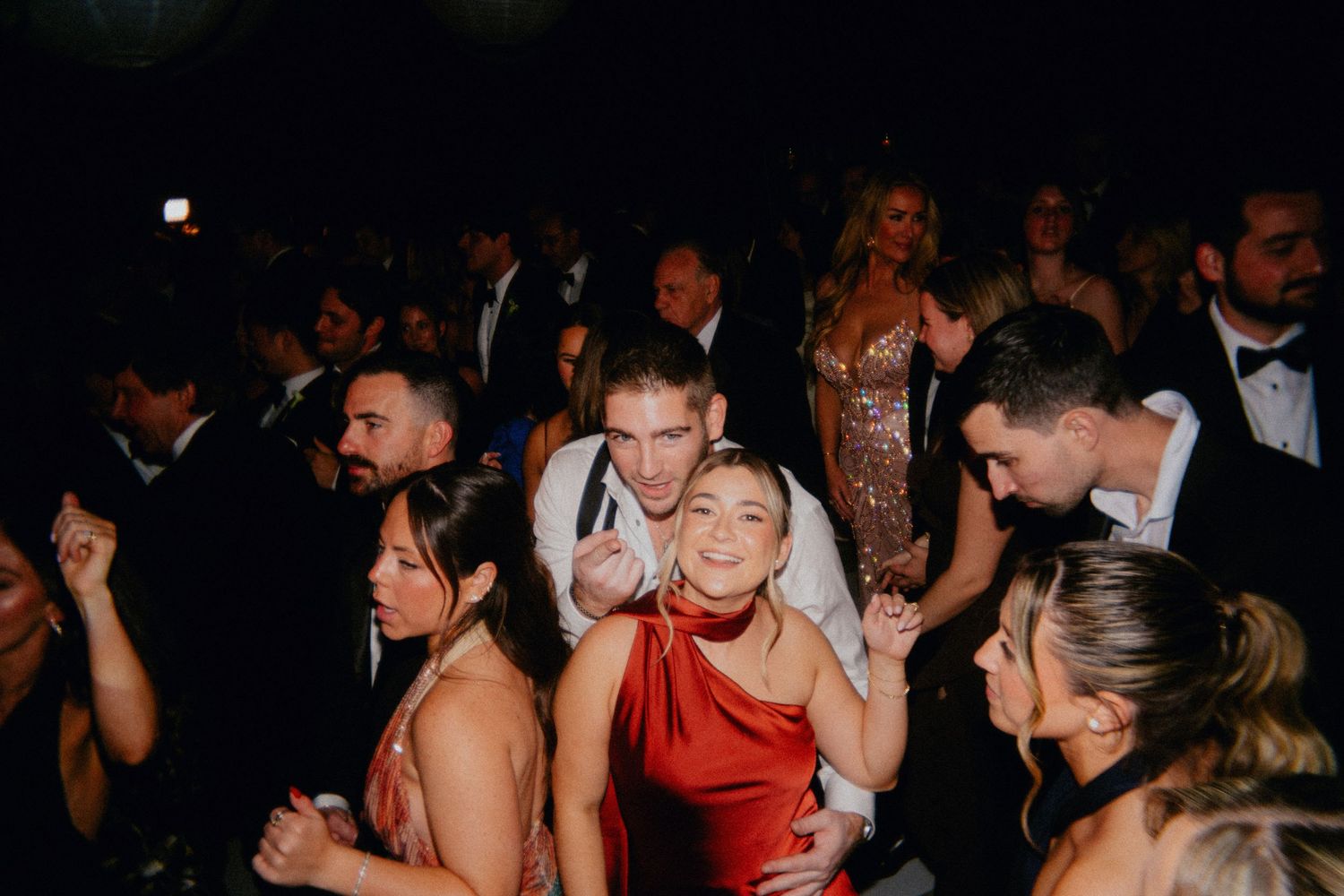 Guests dancing and celebrating at a formal event, wearing elegant attire including red dress and tuxedos.