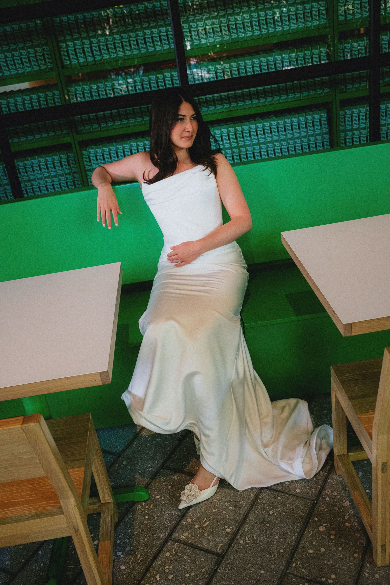 Bride in elegant white satin mermaid gown posing gracefully against a vibrant green wall in a modern venue.