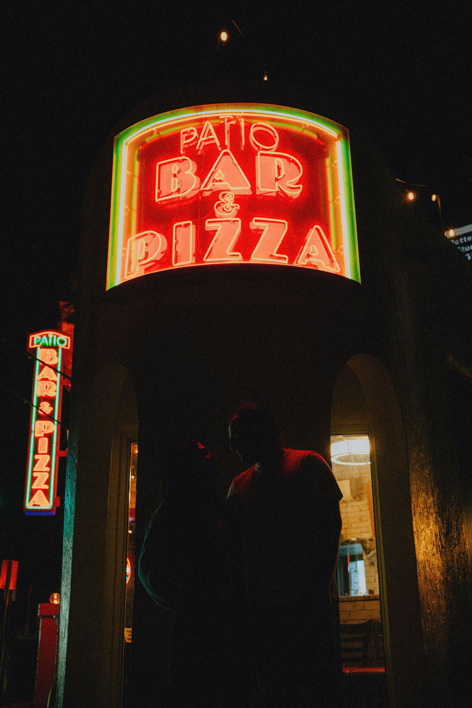 Silhouette of a person standing beneath a glowing neon Patio Bar & Pizza sign at night.