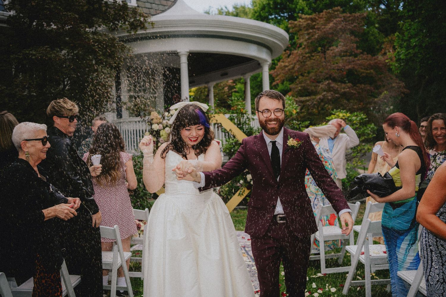 Joyful newlyweds walk down outdoor aisle as guests toss confetti near garden gazebo.