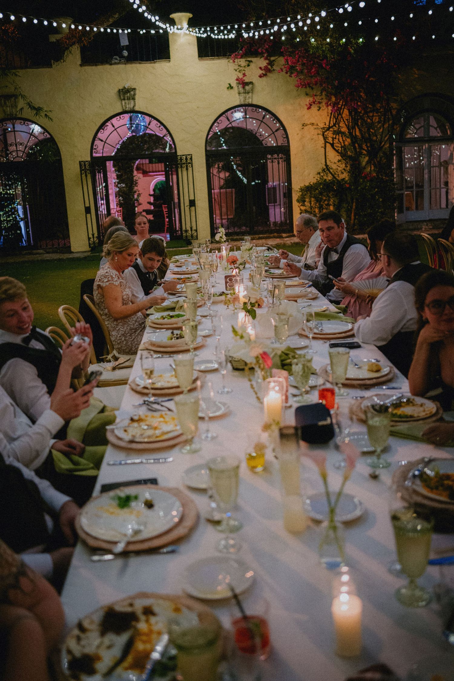 Guests seated at a long candlelit outdoor wedding reception table beneath string lights at an elegant venue.