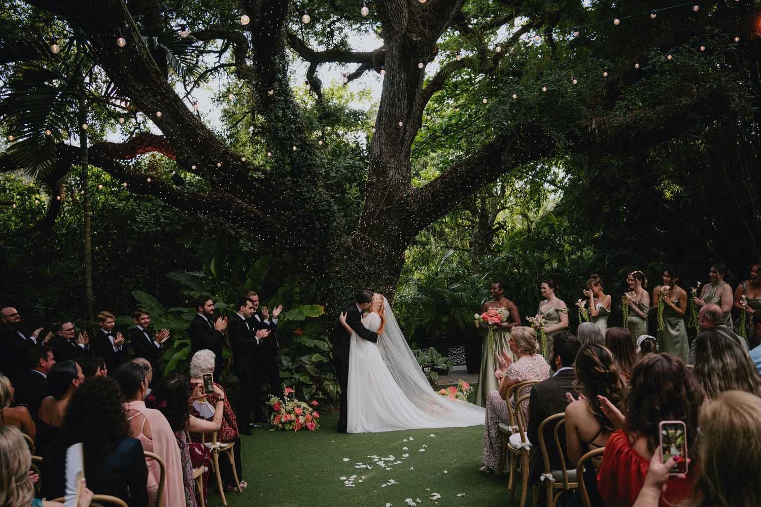 Bride and groom share a kiss at an outdoor wedding ceremony under a large oak tree with string lights.