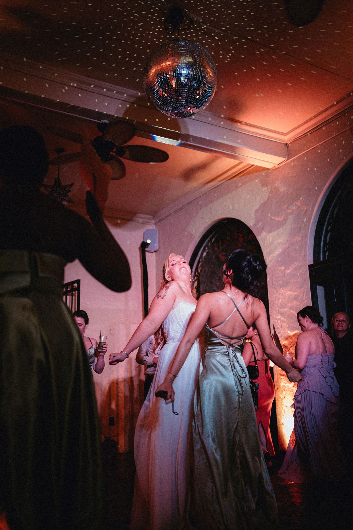 Two brides dance joyfully under a disco ball at their wedding reception in a warmly lit venue.