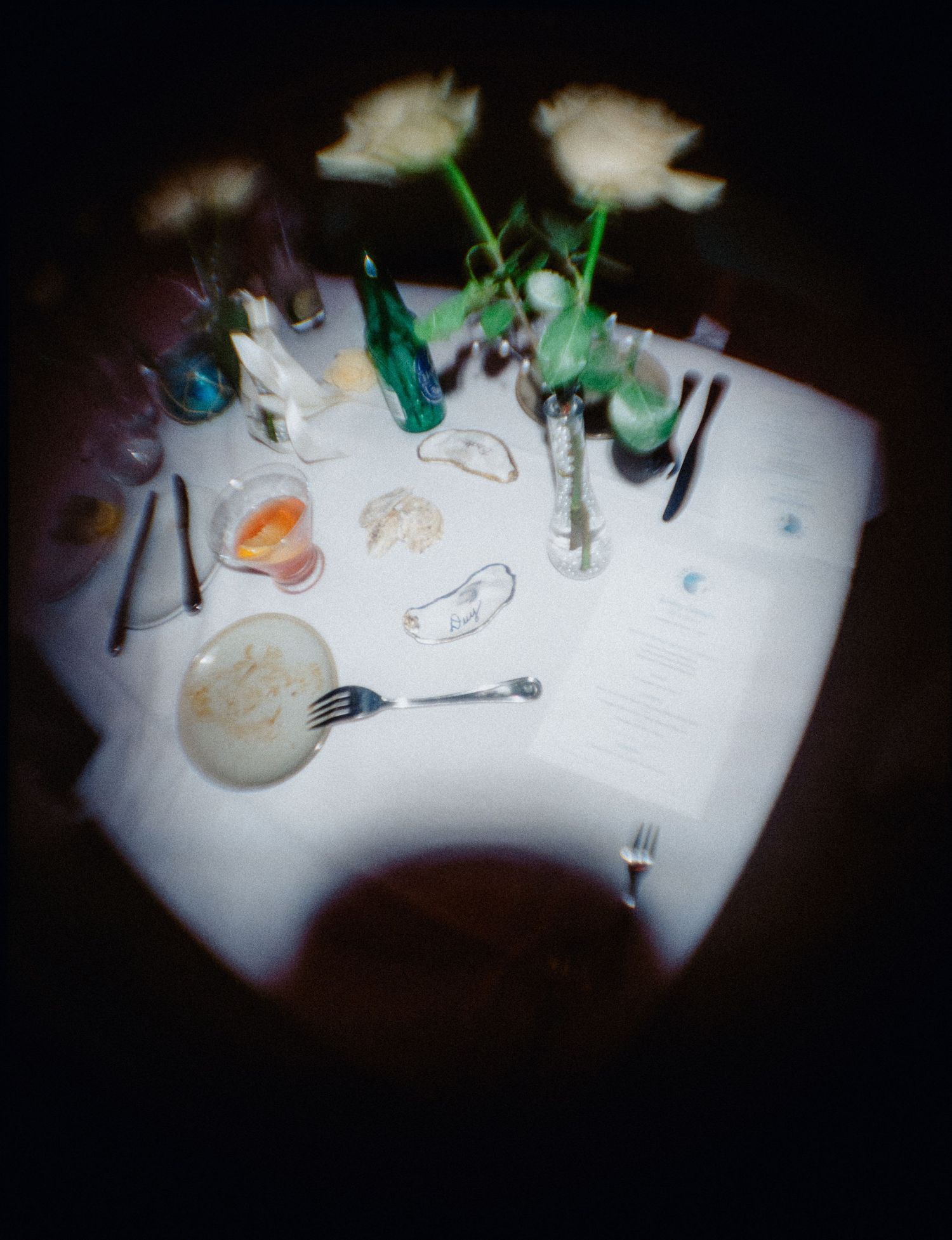 Overhead view of a white-clothed dining table set with white roses in glass vases, a menu card, oyster shells, a fork and knives.