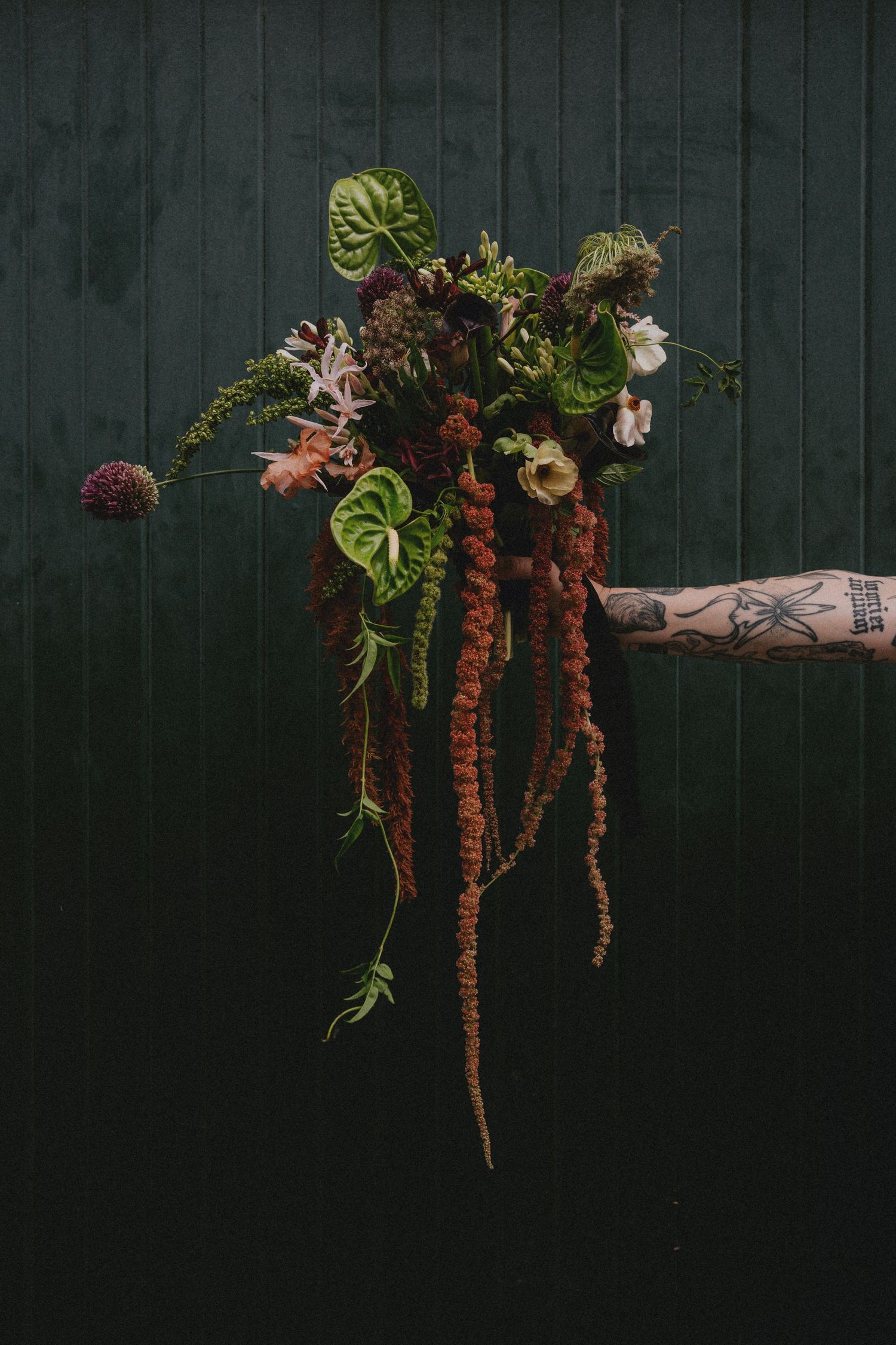 Tattooed hand holding a wild botanical bouquet with ferns, greenery, and flowers against a dark green wall.
