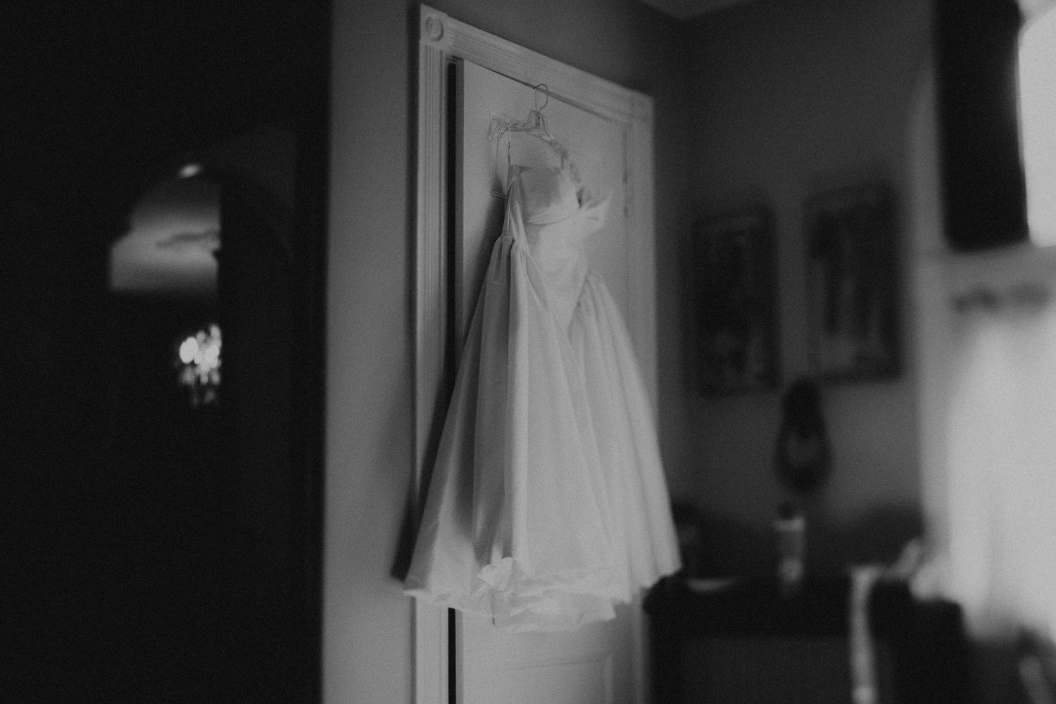 Black and white photo of an elegant wedding dress hanging on a door in a softly lit room.