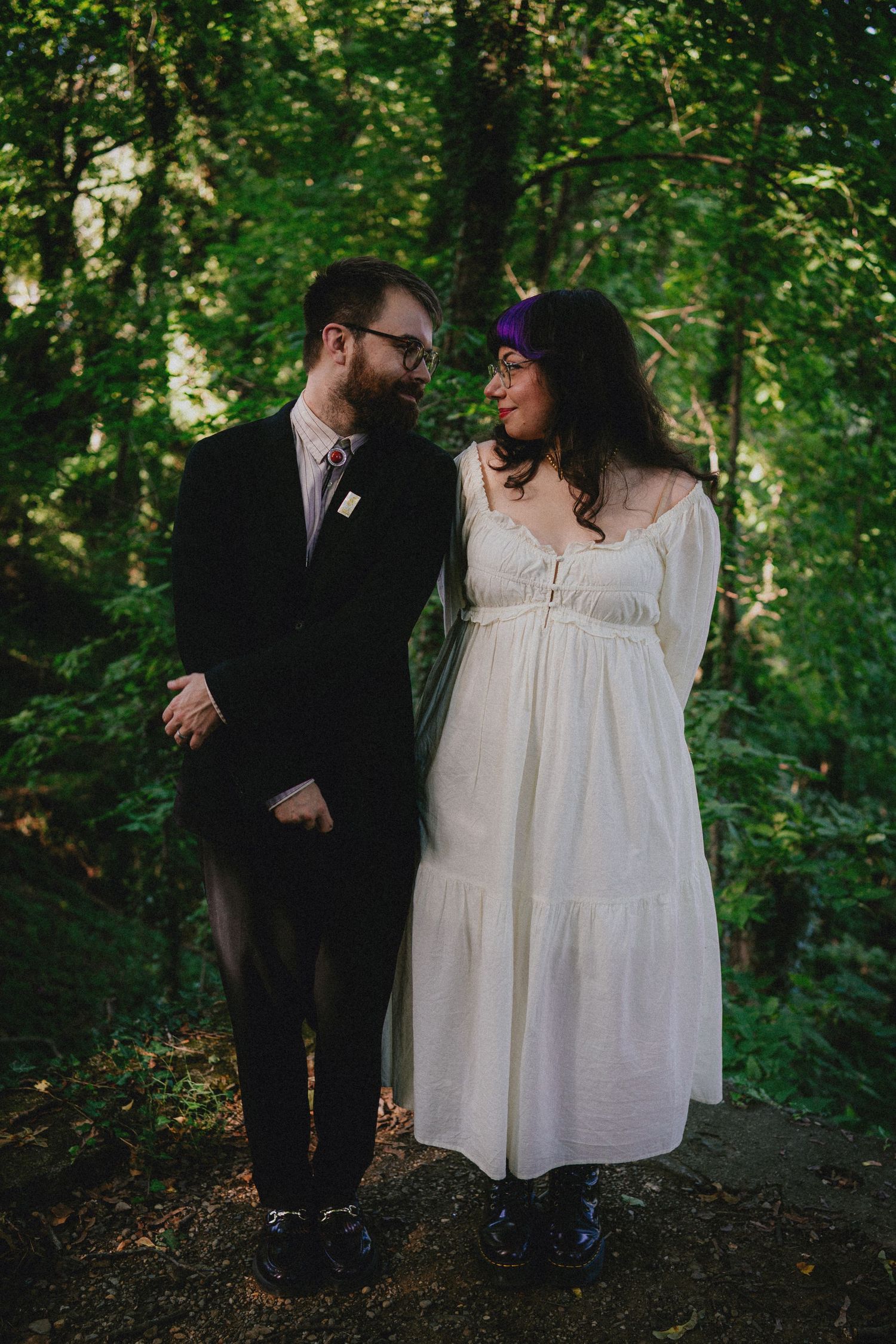 A couple in wedding attire shares a romantic moment in a lush green forest, bride in white dress, groom in black suit.