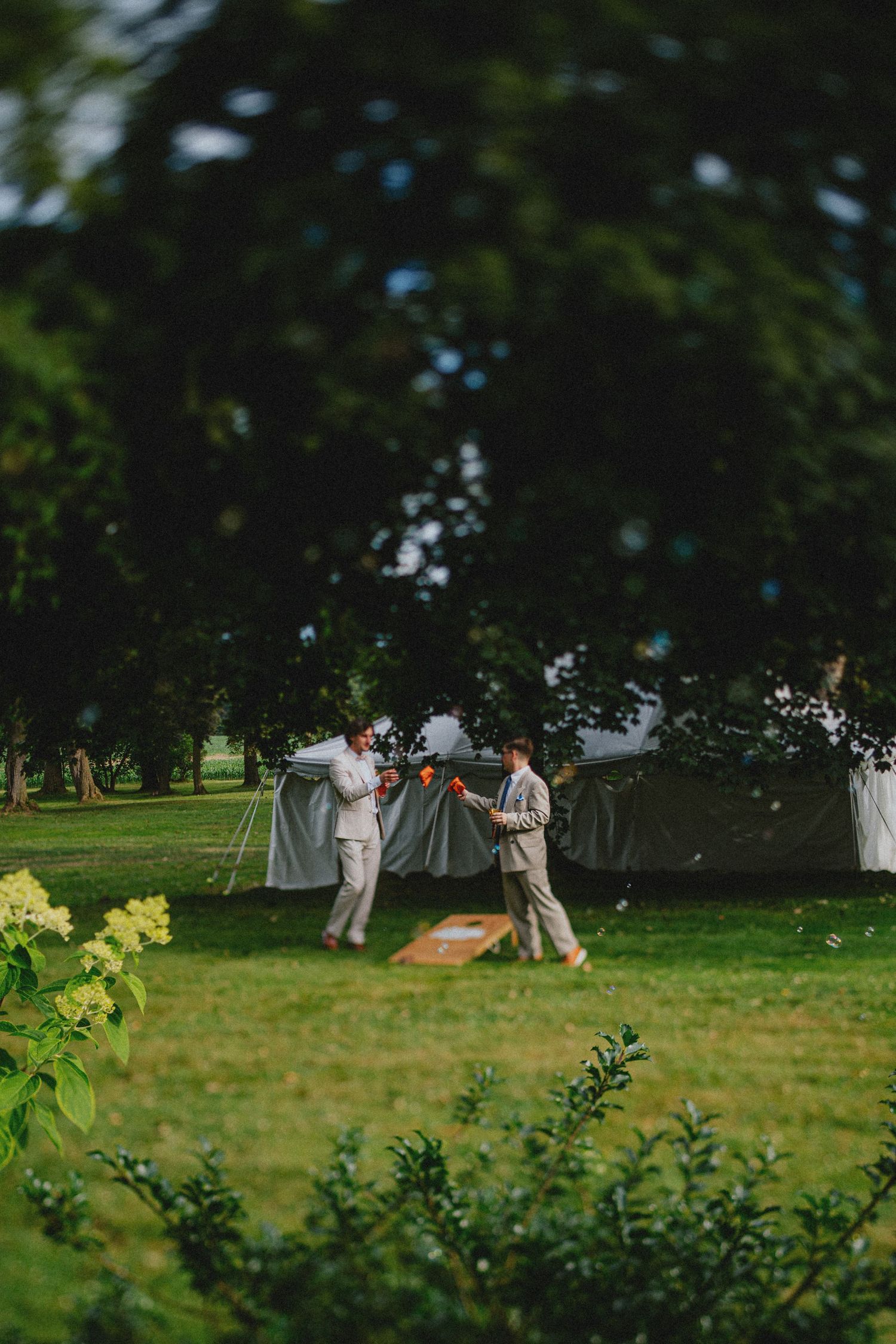 Two groomsmen in tan suits playing cornhole on a green lawn at an outdoor wedding reception.