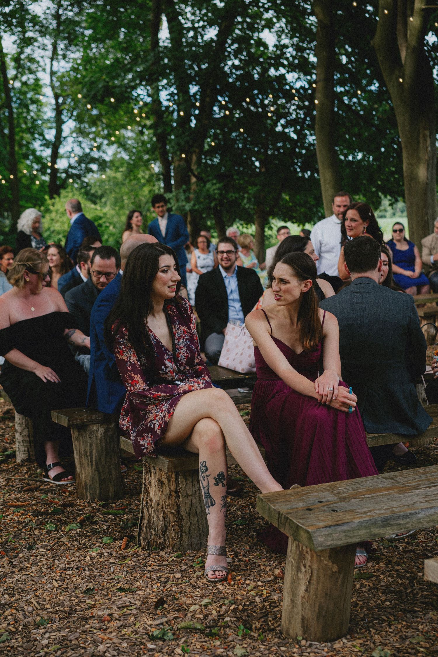 Guests seated on rustic wooden benches at an outdoor woodland wedding ceremony, with women in floral and burgundy dresses.