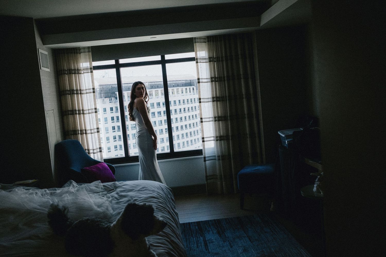 Bride in wedding dress standing by large hotel room window, silhouetted against city skyline view.