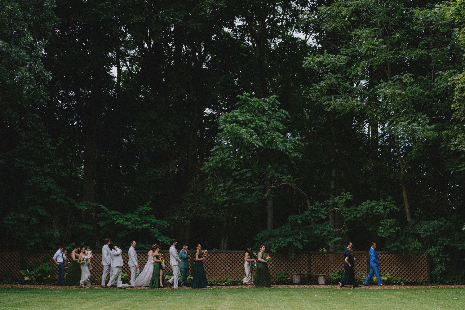Wedding guests gather on a lush green lawn surrounded by tall trees during an outdoor summer ceremony.