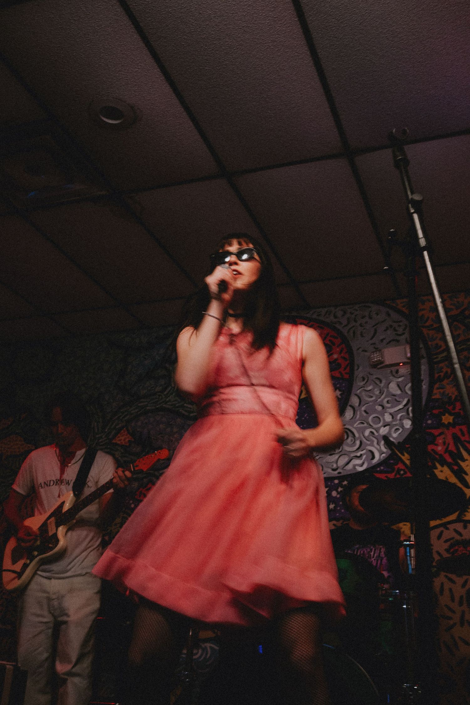 Female vocalist in pink dress performing on stage with guitarist behind her at a live music concert.