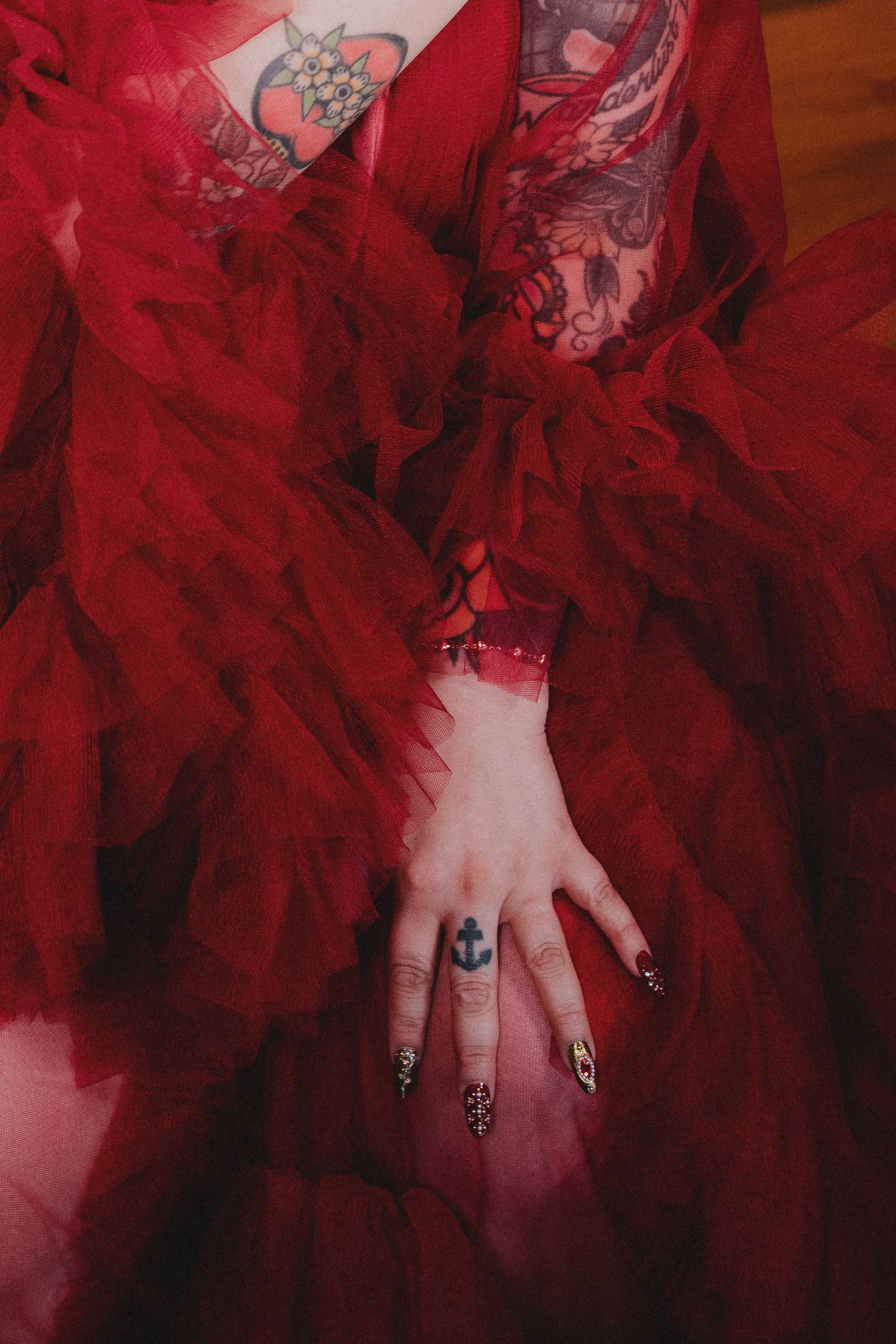 Person in red tulle dress with anchor tattoo on hand, dark nail art, and colorful tattoos visible on arms.