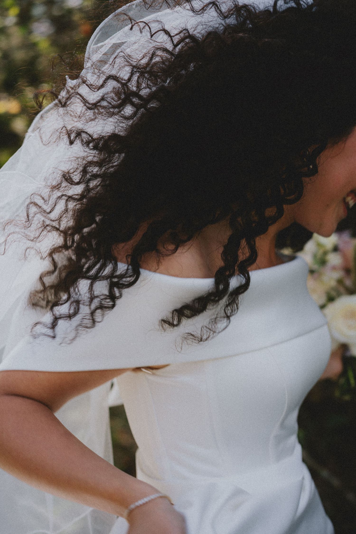Bride with curly dark hair and white wedding dress holding a bouquet, veil flowing in the breeze.