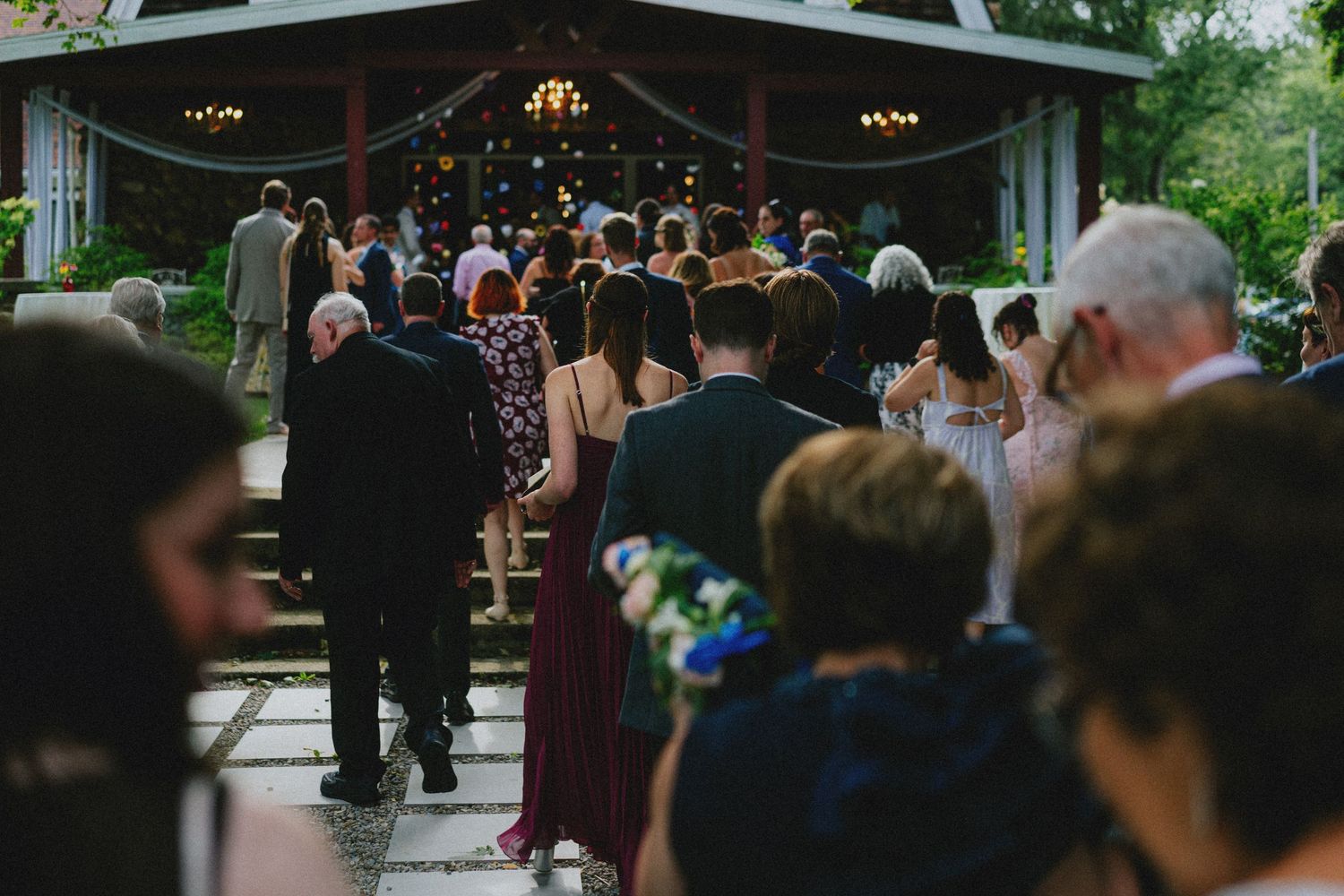 Wedding guests walk toward an outdoor ceremony venue surrounded by trees and string lights.