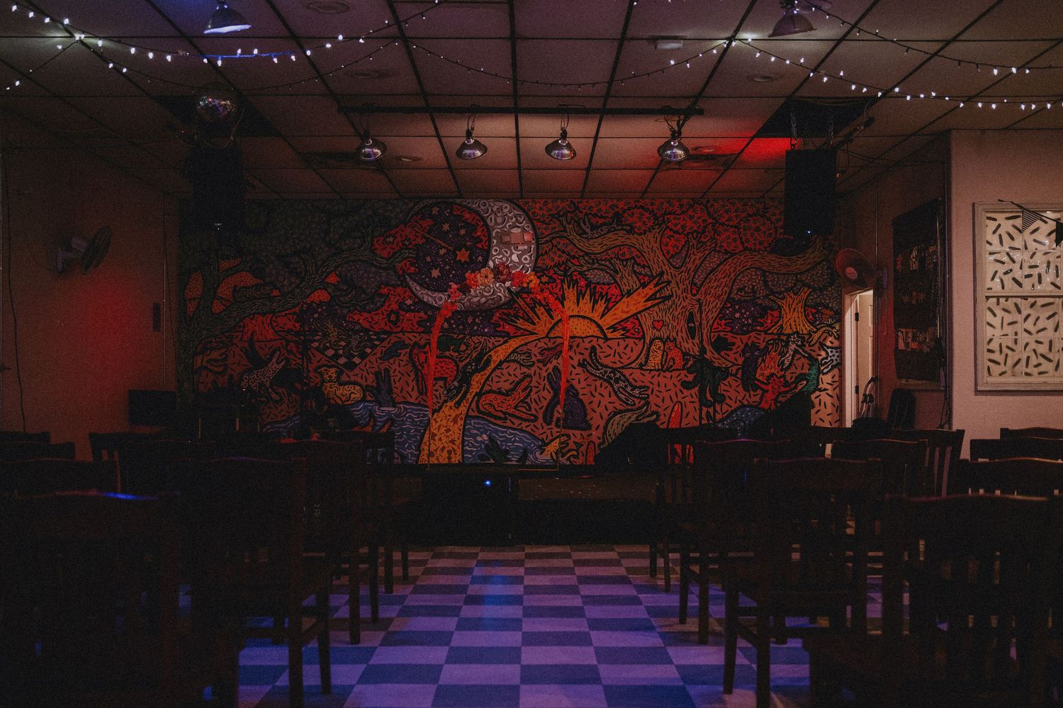 Dark venue with colorful mural, checkered floor, string lights, and rows of chairs in moody red and blue lighting.