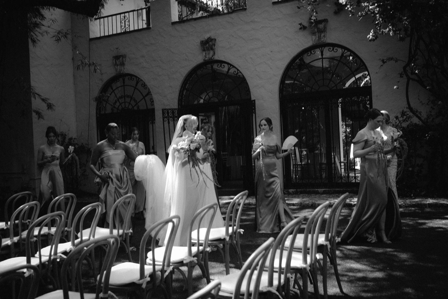 Black and white photo of a bride in white gown with bridesmaids in long dresses walking through outdoor wedding venue.