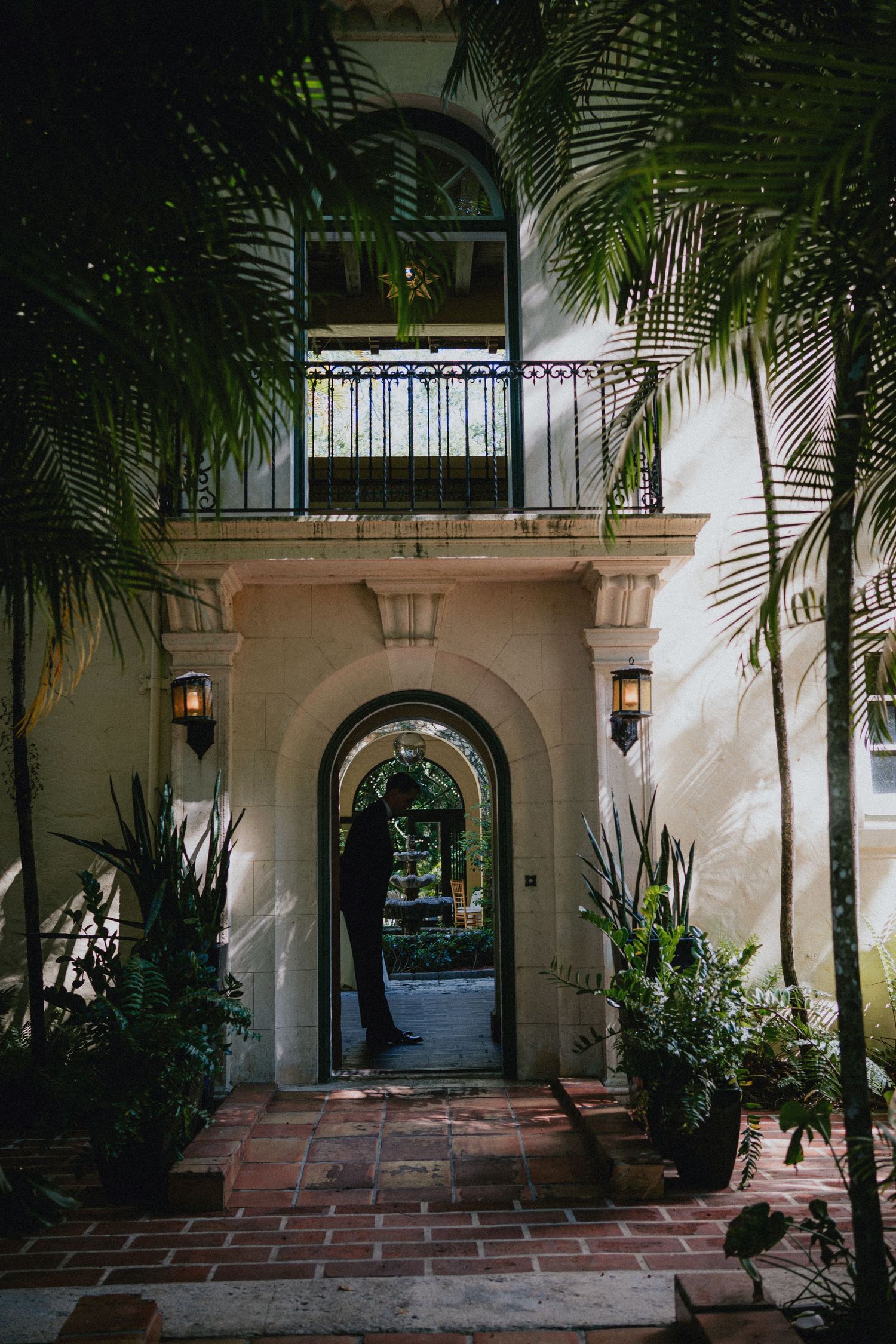 Elegant Mediterranean villa entrance with arched doorway, wrought iron balcony, palm trees, and brick pathway at dusk.