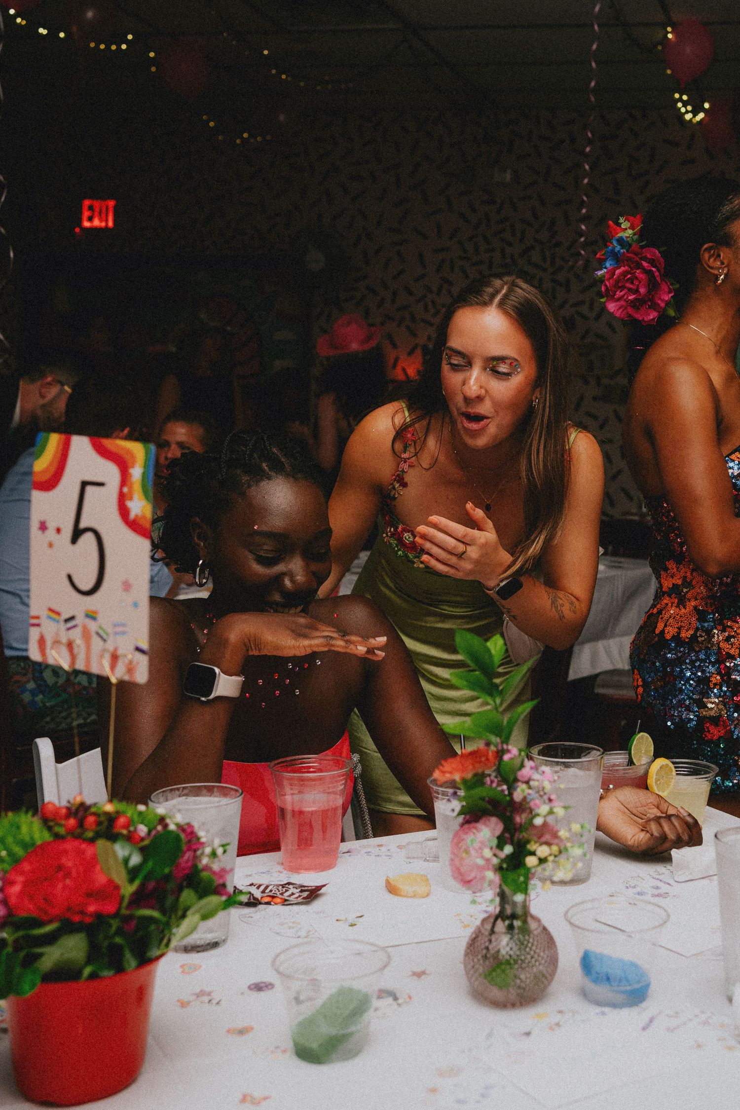 Two women celebrate at a colorful party, leaning over a decorated table with flowers and drinks at table number 5.